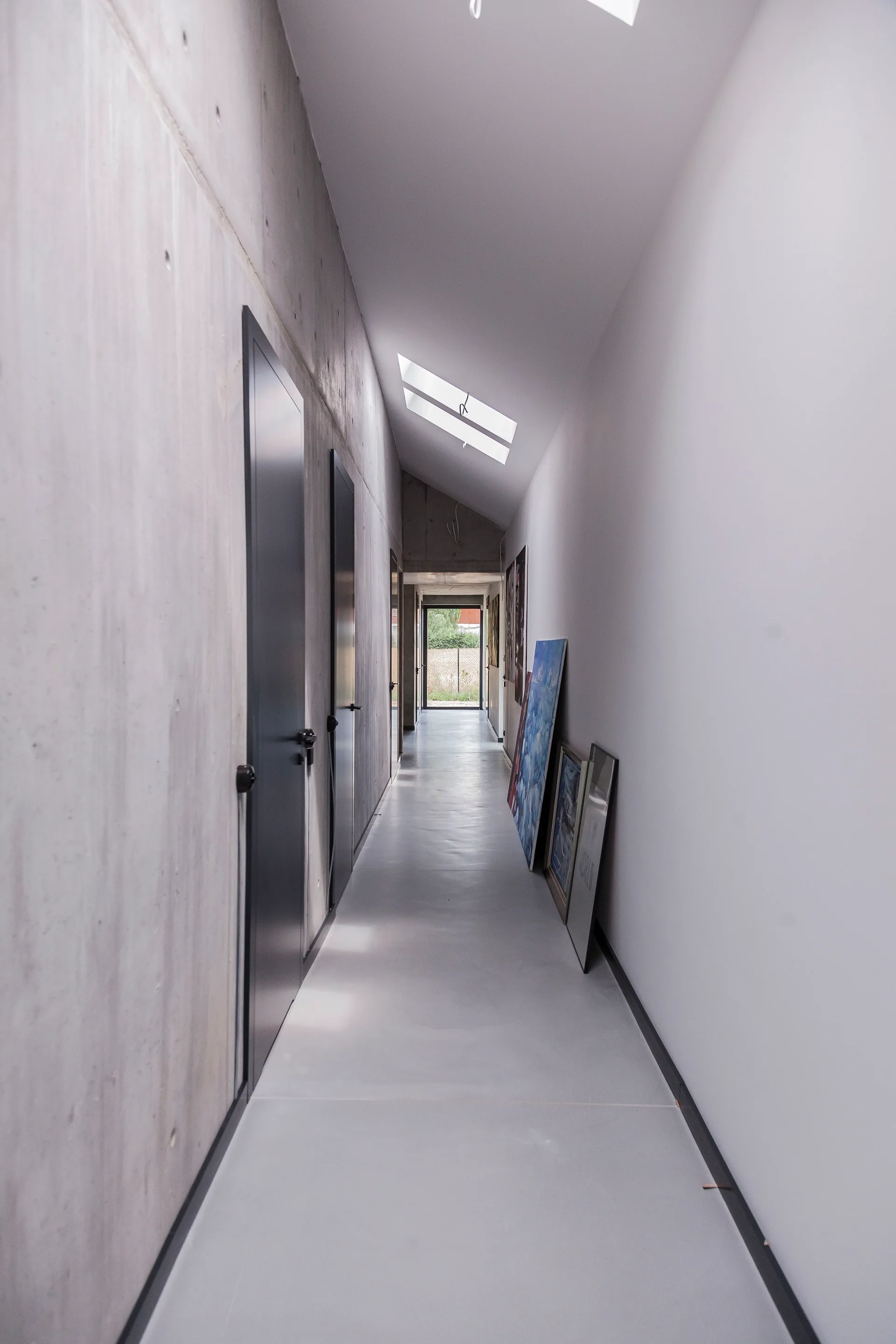 A minimalist hallway with an exposed concrete wall, dark flush doors, sloped skylights, and framed art leaning against a white wall.