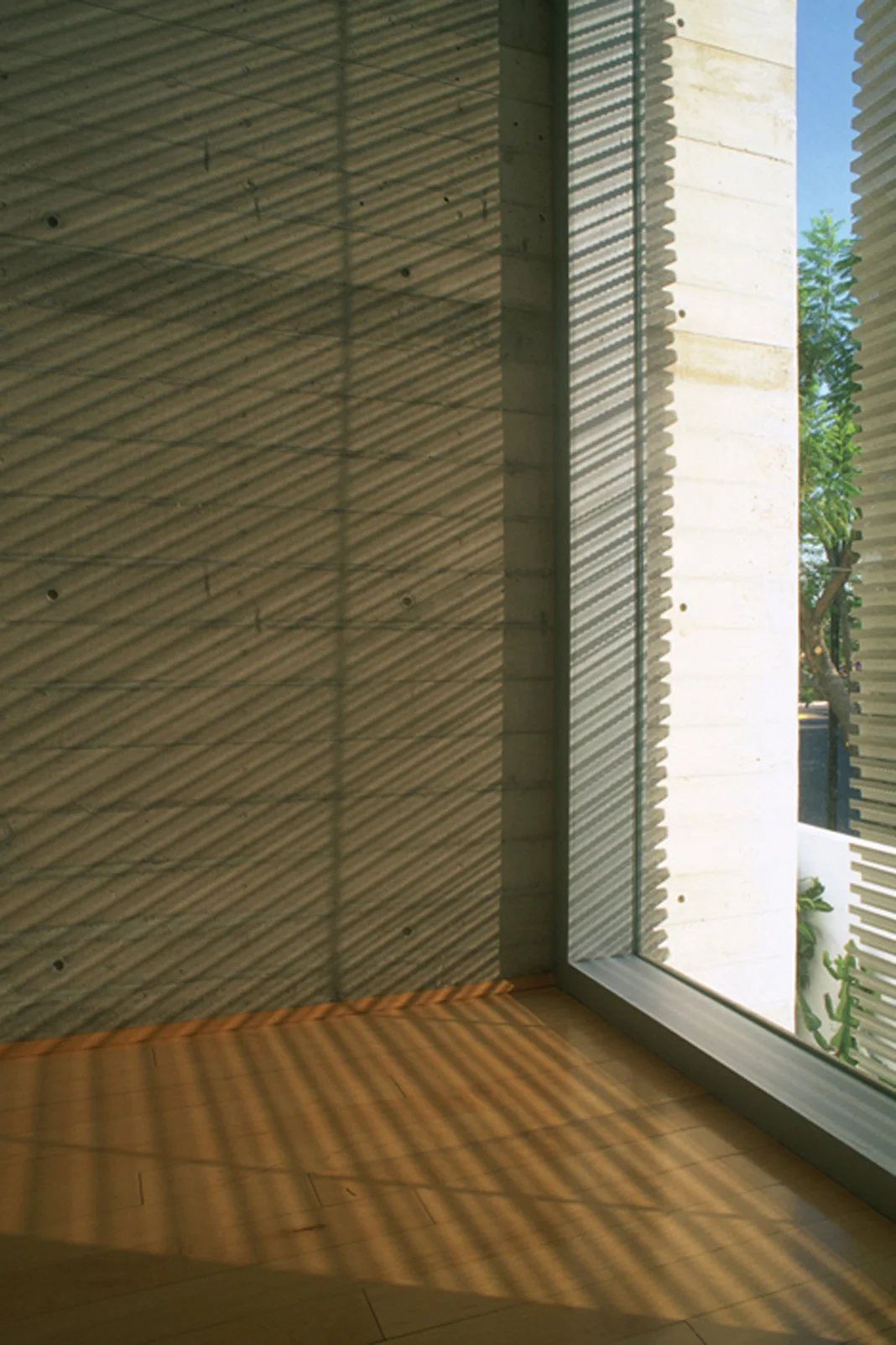 Diagonal striped shadows from window blinds fall across a raw concrete wall and light hardwood flooring in a minimalist interior.