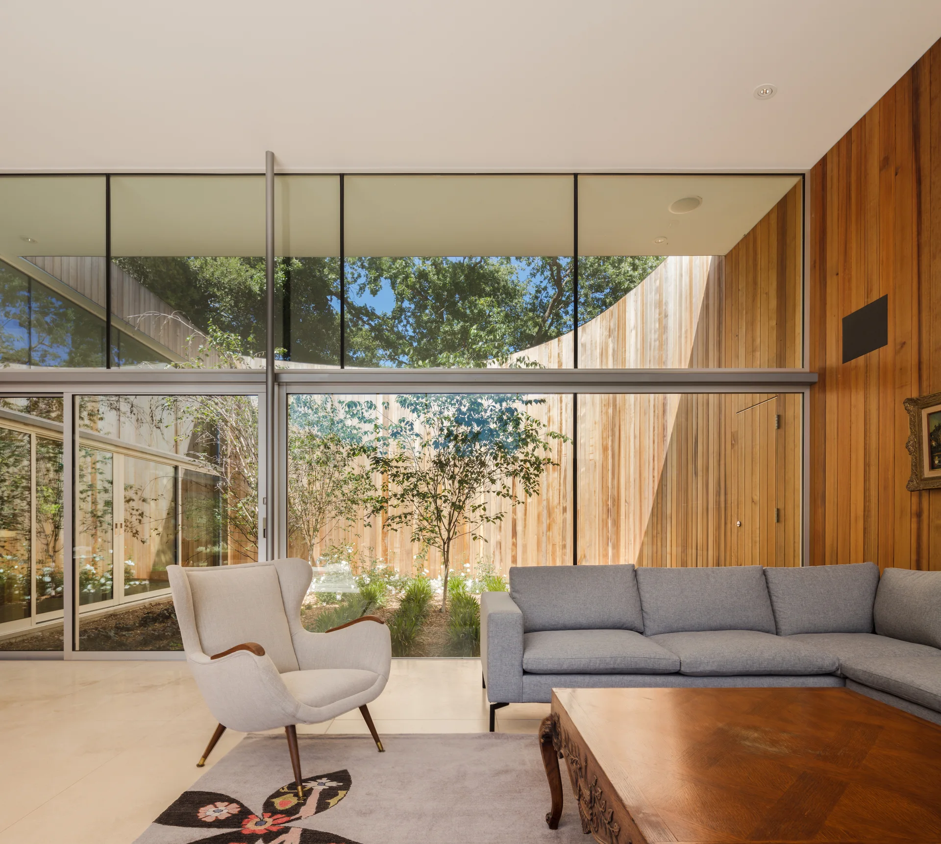 A modern living room with a grey sectional sofa, mid-century armchair, and a large glass wall looking out to a wood-walled courtyard.