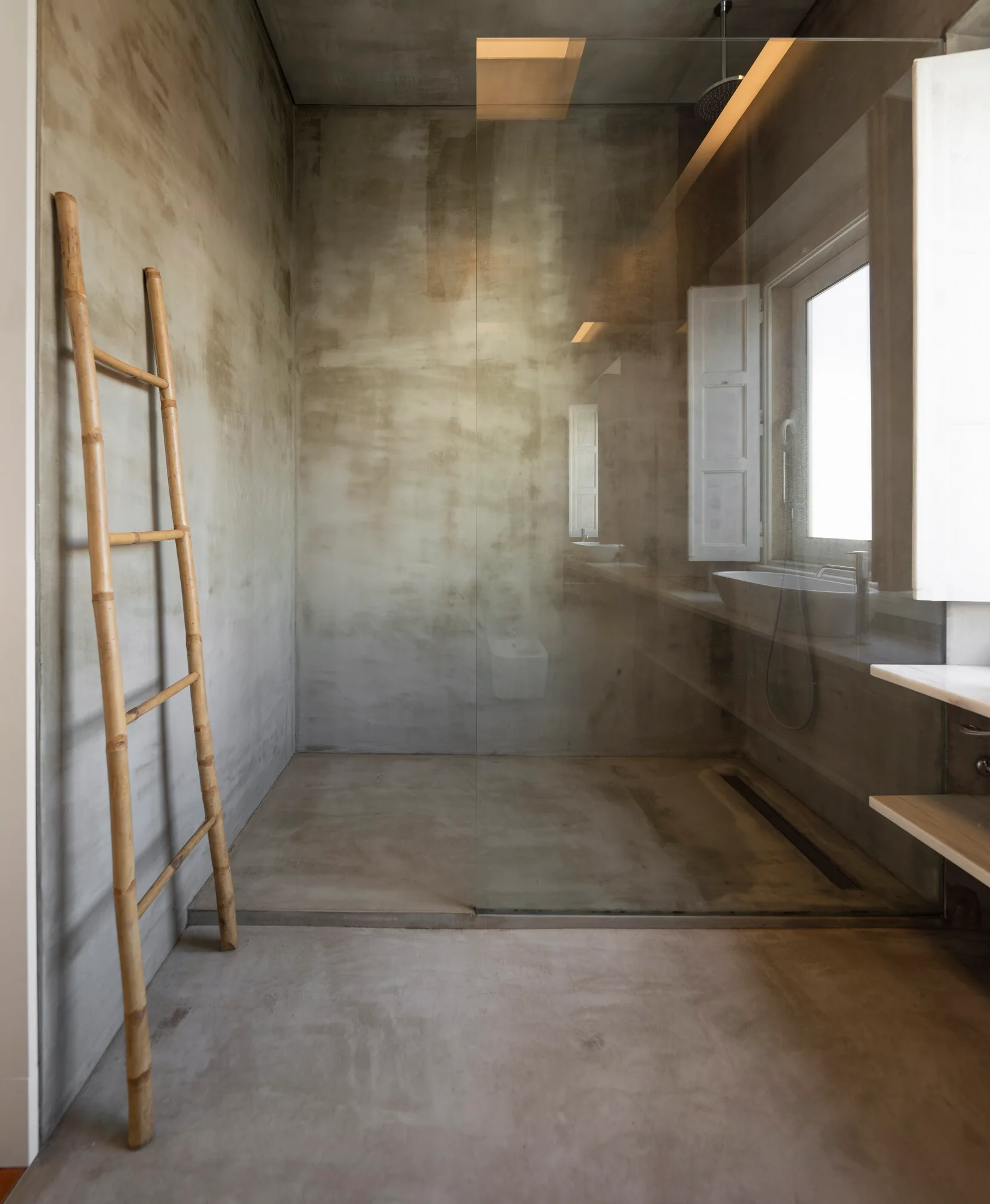 A minimalist bathroom with textured grey concrete walls and flooring featuring a glass shower partition and a leaning bamboo ladder.