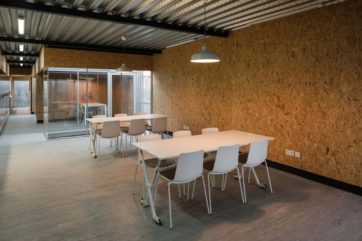 An industrial office space features raw oriented strand board walls, a corrugated metal ceiling, and white tables with chairs.