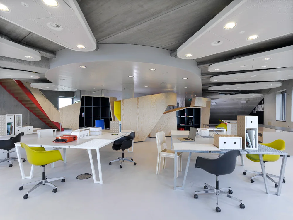 A contemporary open-plan office space featuring white desks with black and lime green chairs, angular plywood partitions, and suspended ceiling panels.