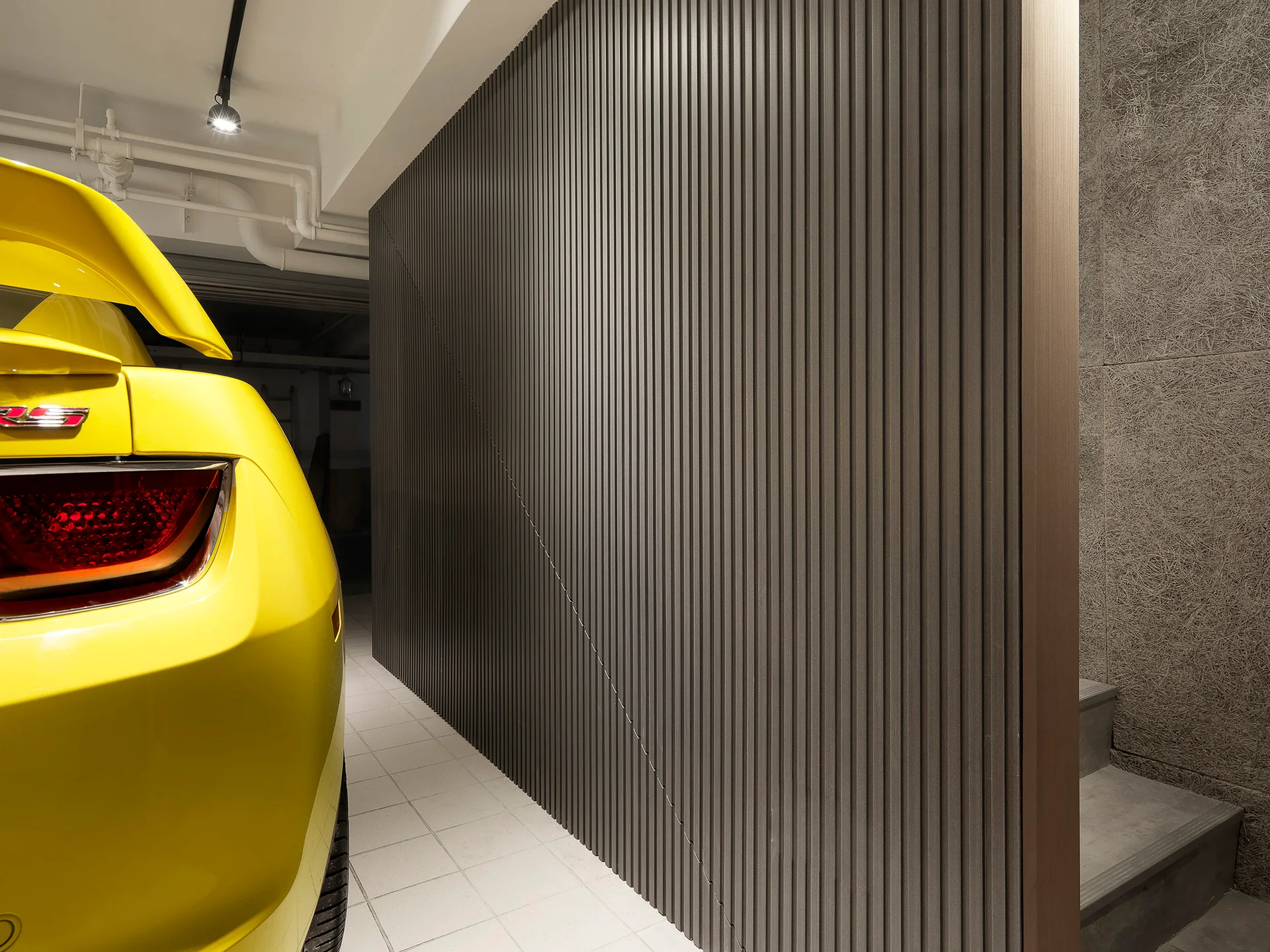 A bright yellow car parked in a garage next to a dark grey slatted wall, white tiled floors, and a concrete staircase.
