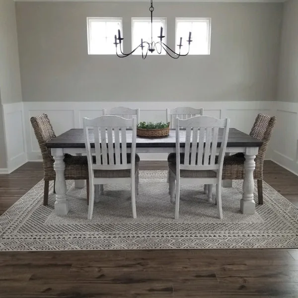 A neutral-toned dining room features a dark-top farmhouse table surrounded by white wooden and woven rattan chairs on a geometric patterned rug.