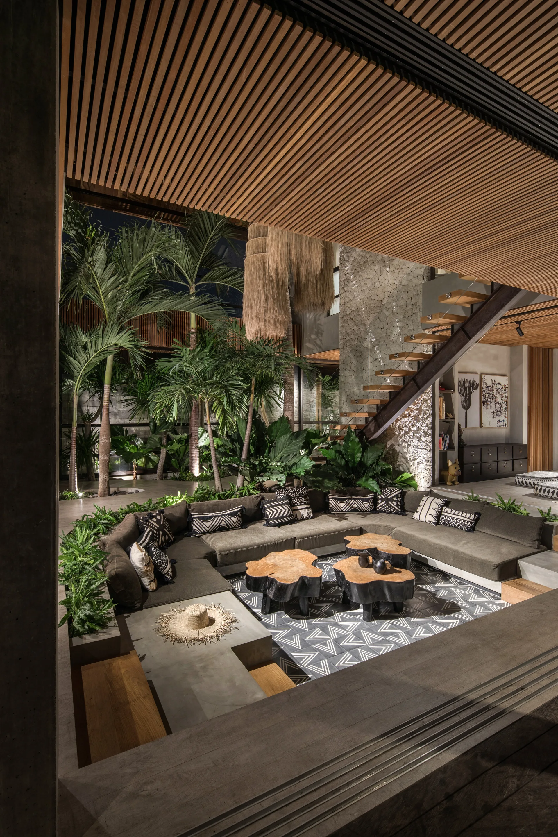 A sunken living room features a dark grey sectional sofa, live-edge wood coffee tables, a slatted timber ceiling, and a lush indoor palm garden.