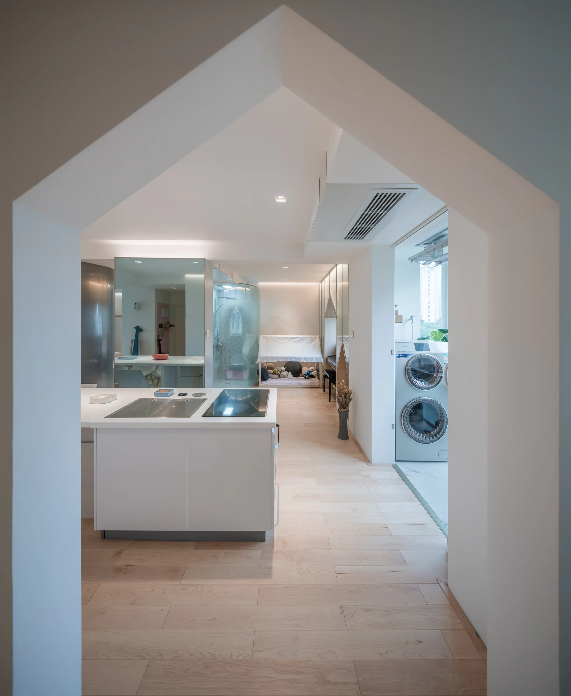 A view through a house-shaped white archway looking into a minimalist kitchen with a white island and light wood flooring.
