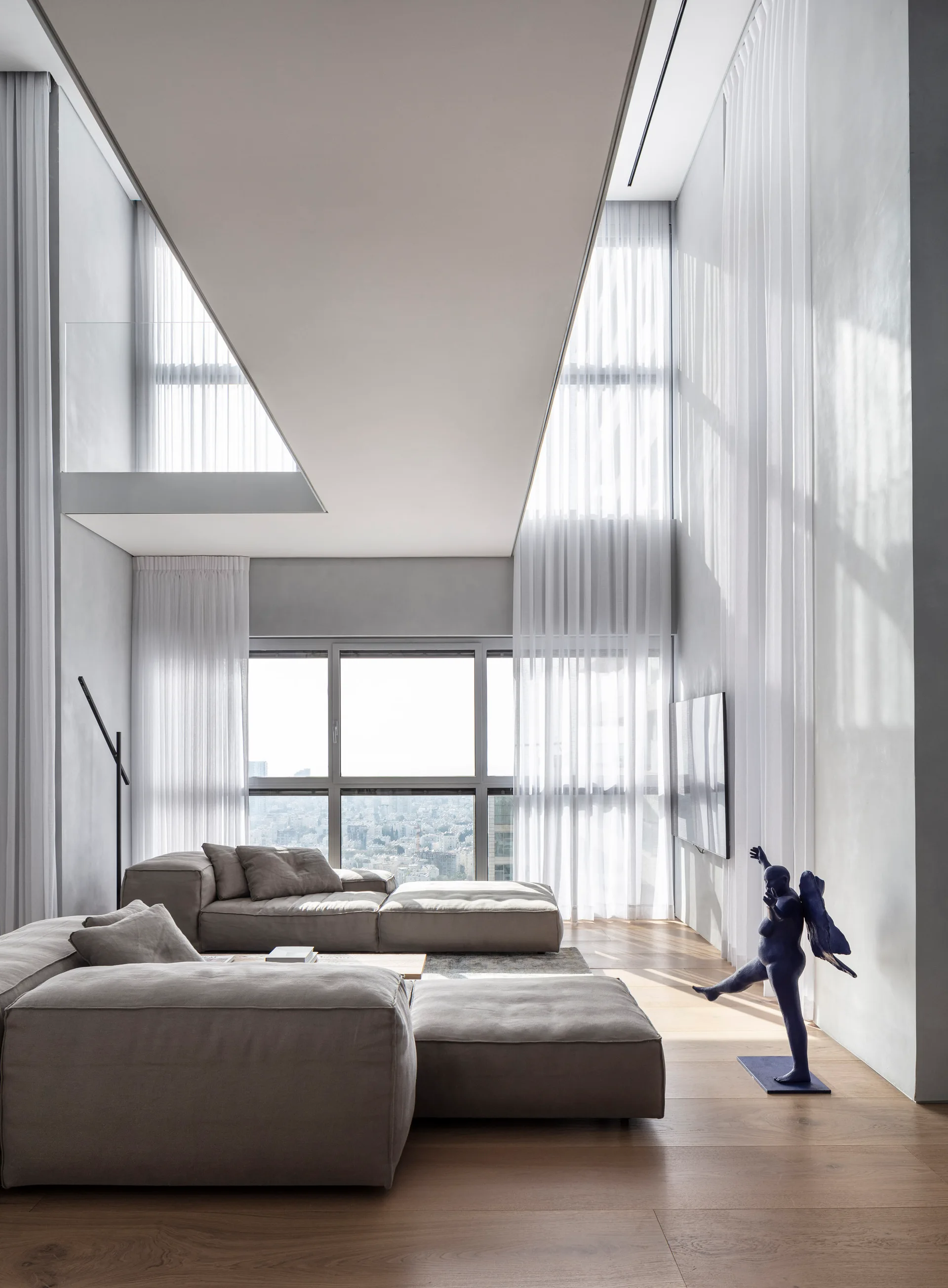 A minimalist double-height living room features a gray modular sofa, floor-to-ceiling windows with sheer white curtains, and wood flooring.