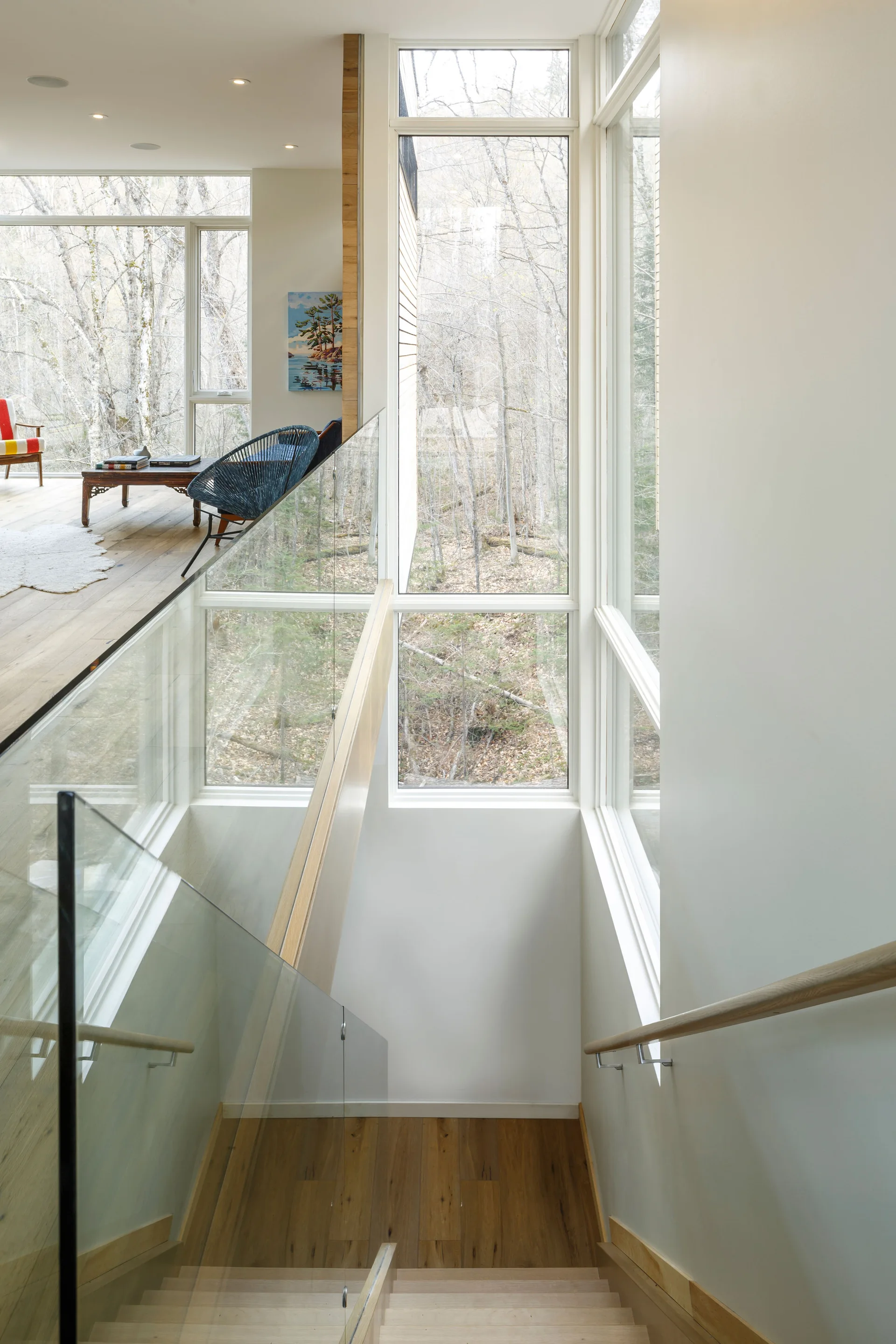 A modern wooden staircase with a transparent glass railing leads down past large windows showcasing a winter forest view.