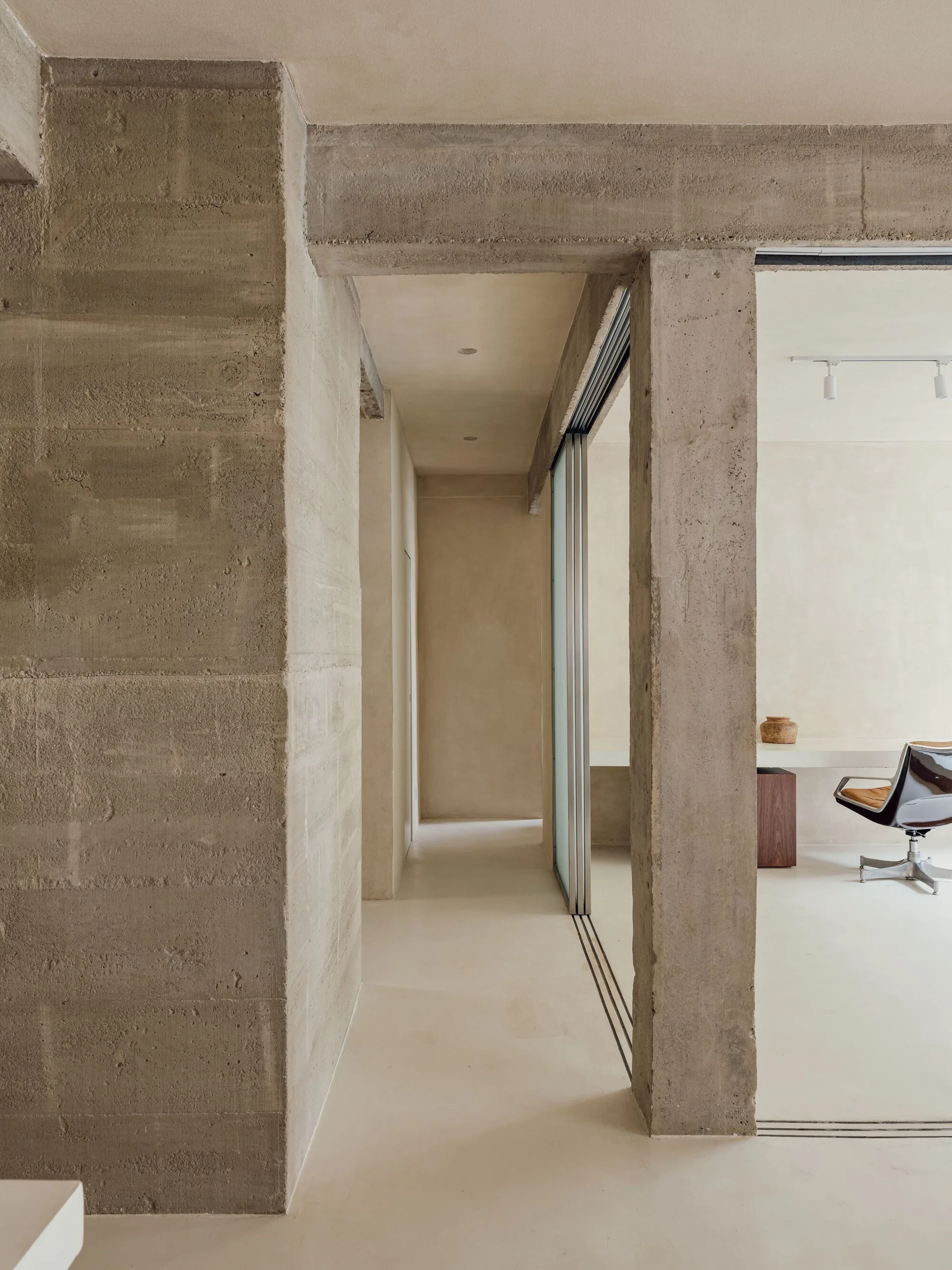 An interior hallway features raw concrete structural pillars leading to a minimalist home office with a white desk, leather chair, and sliding glass partition.