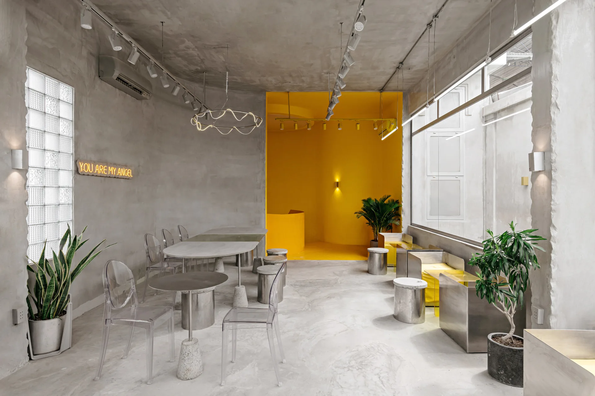 A modern cafe interior with raw concrete walls, transparent acrylic chairs, concrete tables, and a vibrant yellow accent alcove in the background.