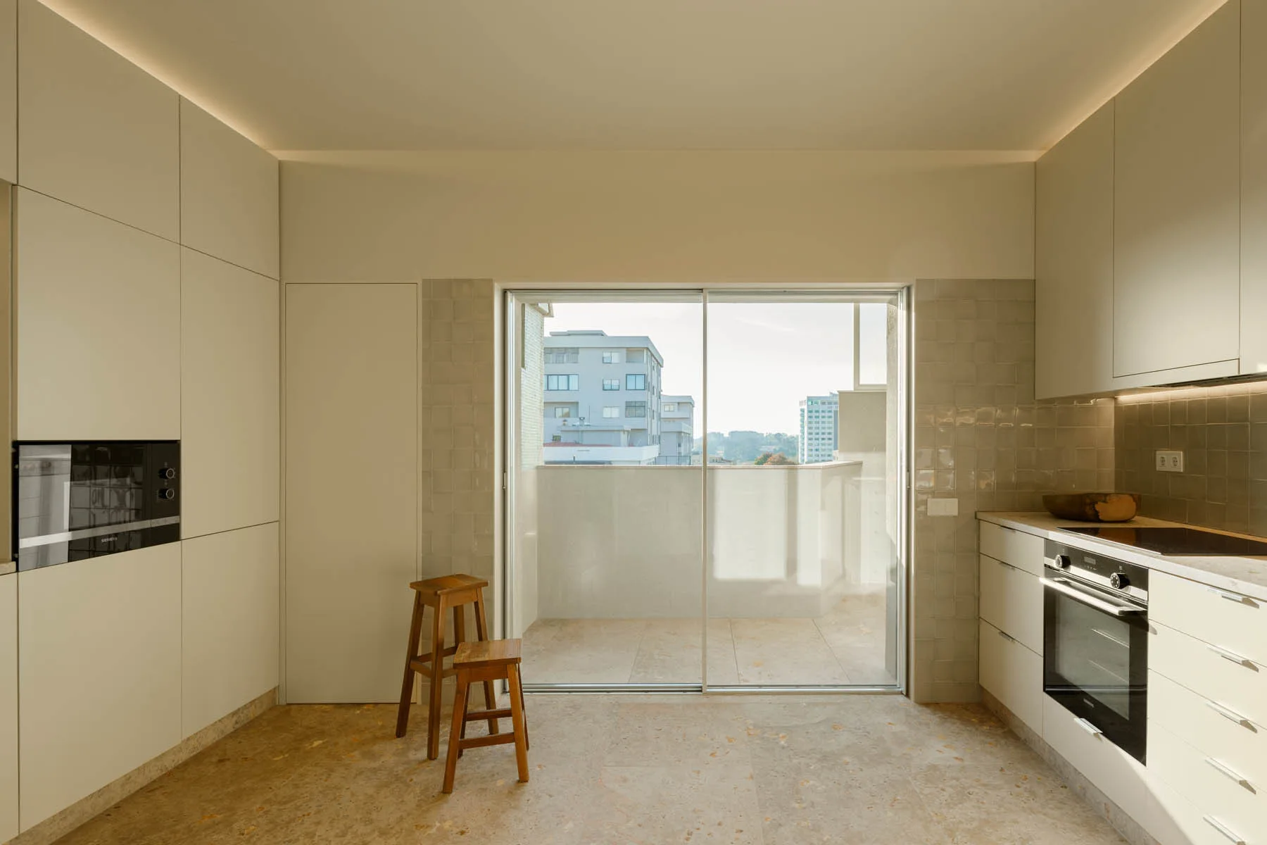 A minimalist kitchen features flat-front off-white cabinets, a built-in oven, two wooden stools, and a sliding glass door leading to a balcony.