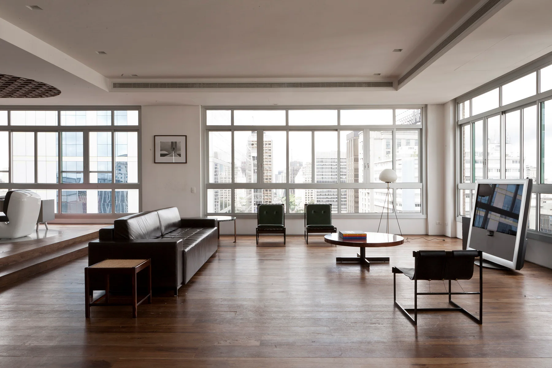 A minimalist living room features a black leather sofa, wooden coffee table, and low-profile chairs arranged on hardwood floors facing large windows.