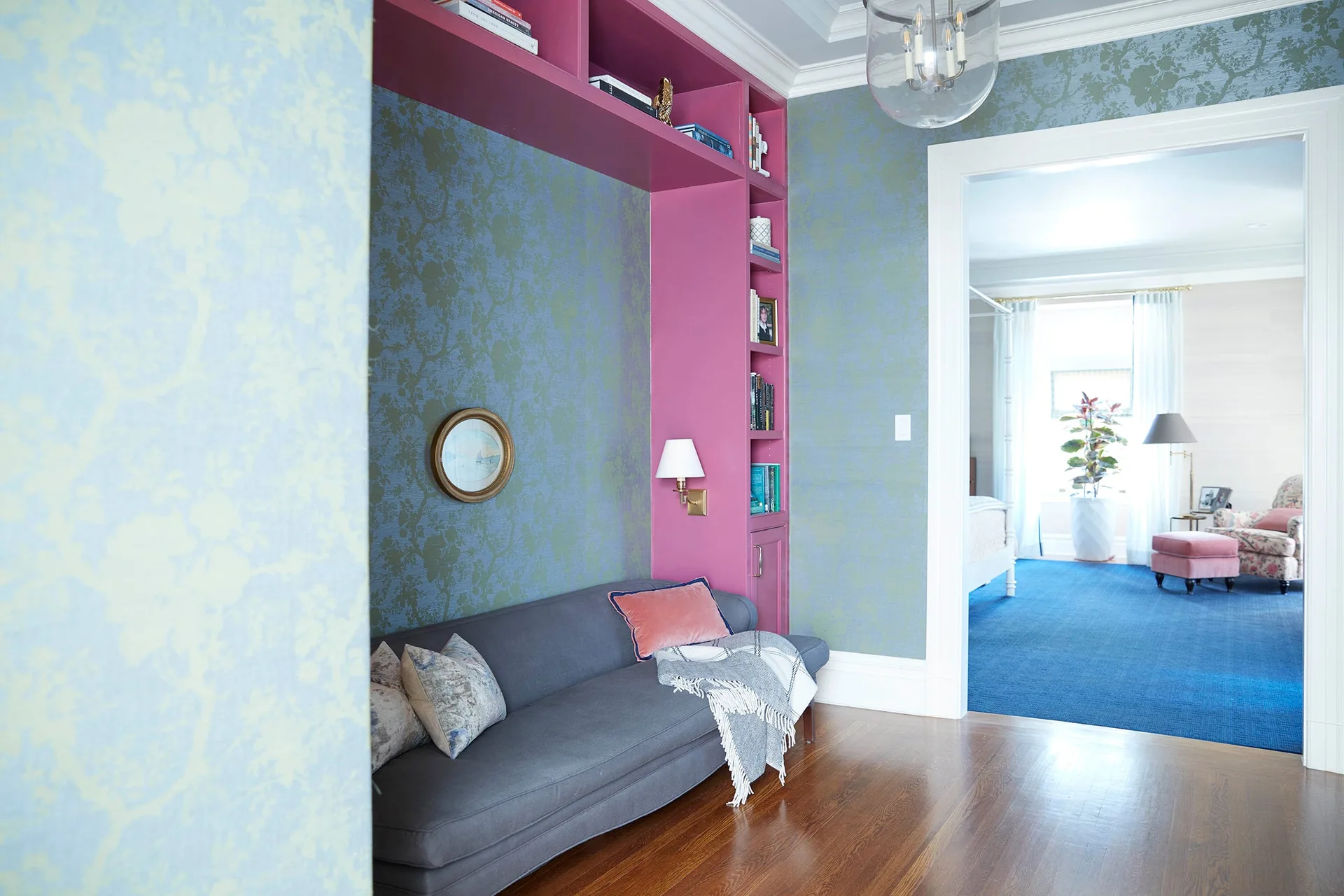 A dark gray sofa sits inside a custom magenta built-in shelving unit on a wall with blue floral wallpaper.