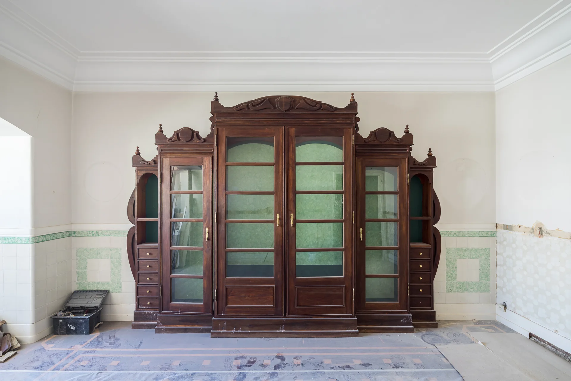 A large ornate dark wood display cabinet with glass doors stands against a white wall with a decorative green patterned tile border.
