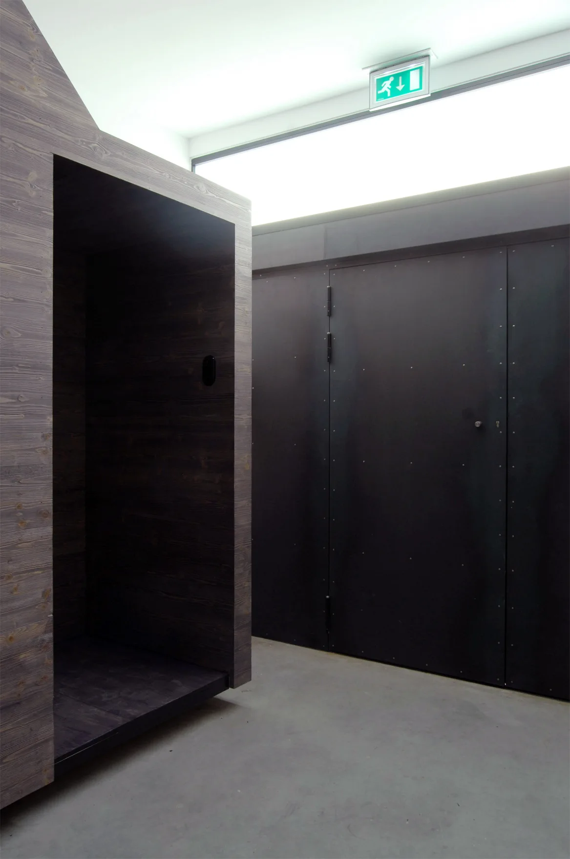 An industrial hallway features a dark wood-grained niche alongside matte black wall panels, concrete flooring, and a glowing green exit sign.