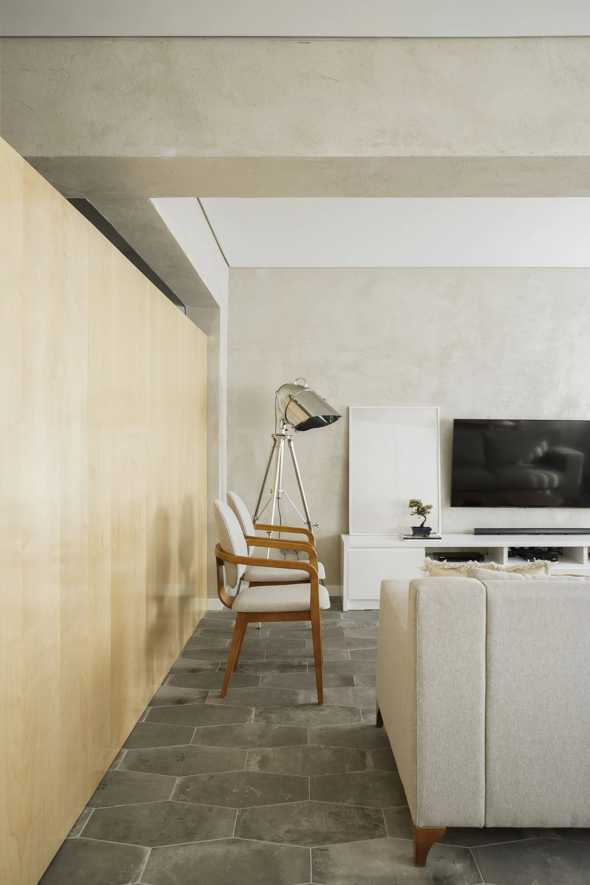 A minimalist living room featuring a light wood paneled wall, wood-framed armchairs, gray stone floor tiles, and a white media console.