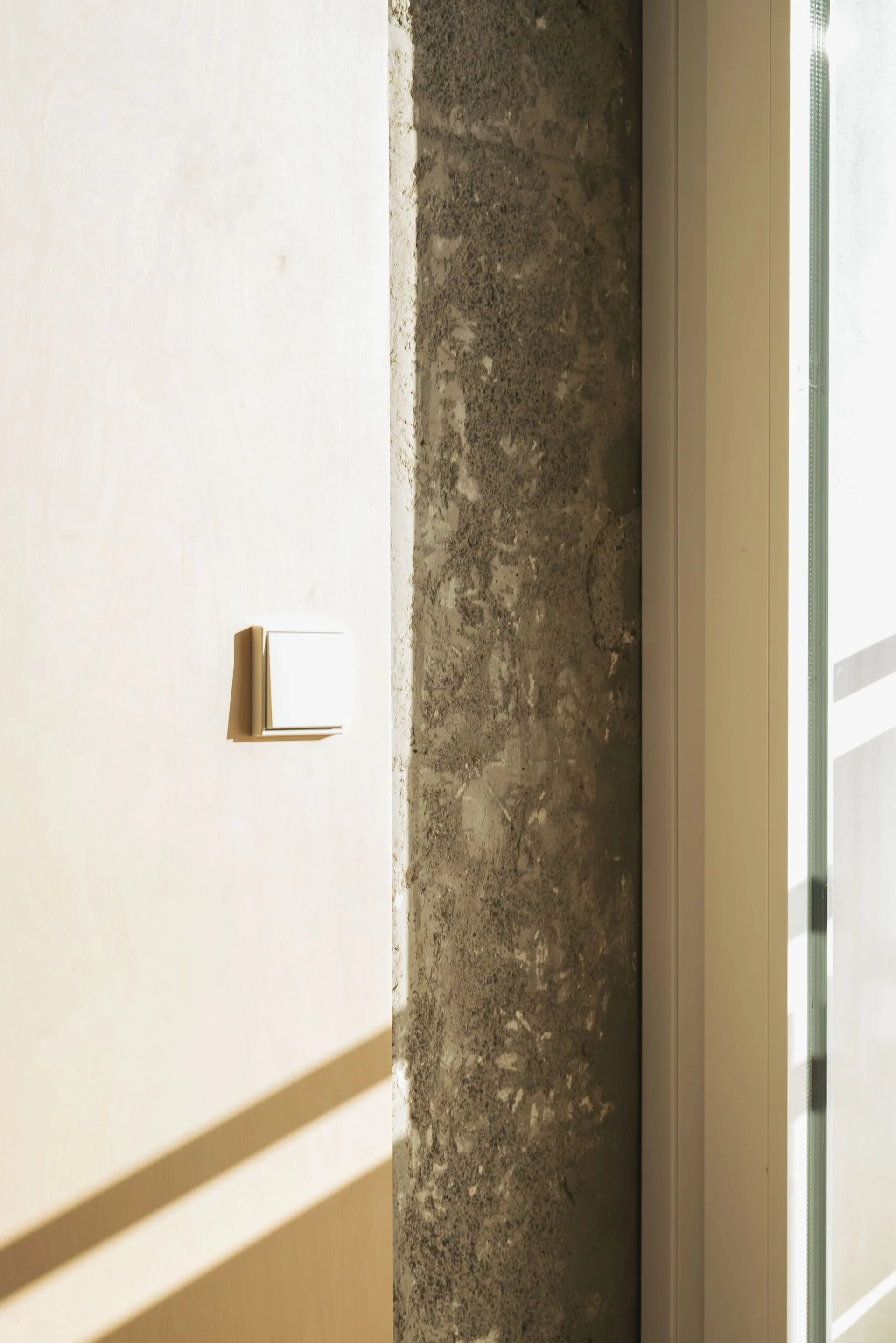 A white square light switch mounted on a light plywood wall panel next to a rough gray concrete pillar.