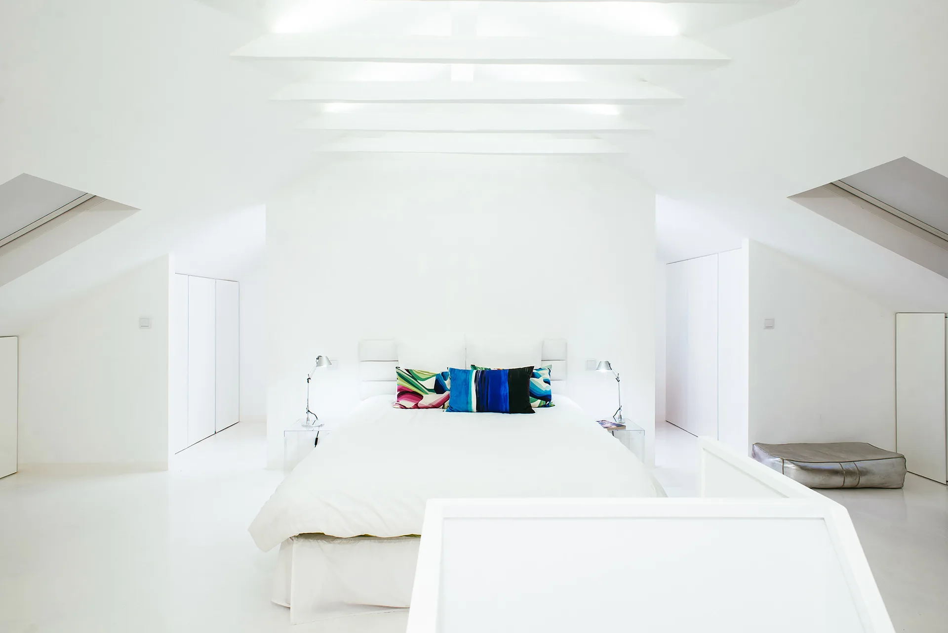 A minimalist white attic bedroom featuring a centered bed with colorful patterned pillows, sloped ceilings, and modern bedside lamps.