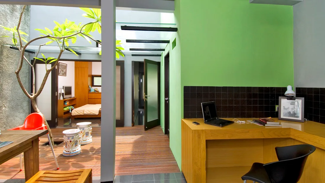 A modern home office features a wooden built-in desk beside a green wall, adjacent to a wooden deck walkway under a glass skylight.