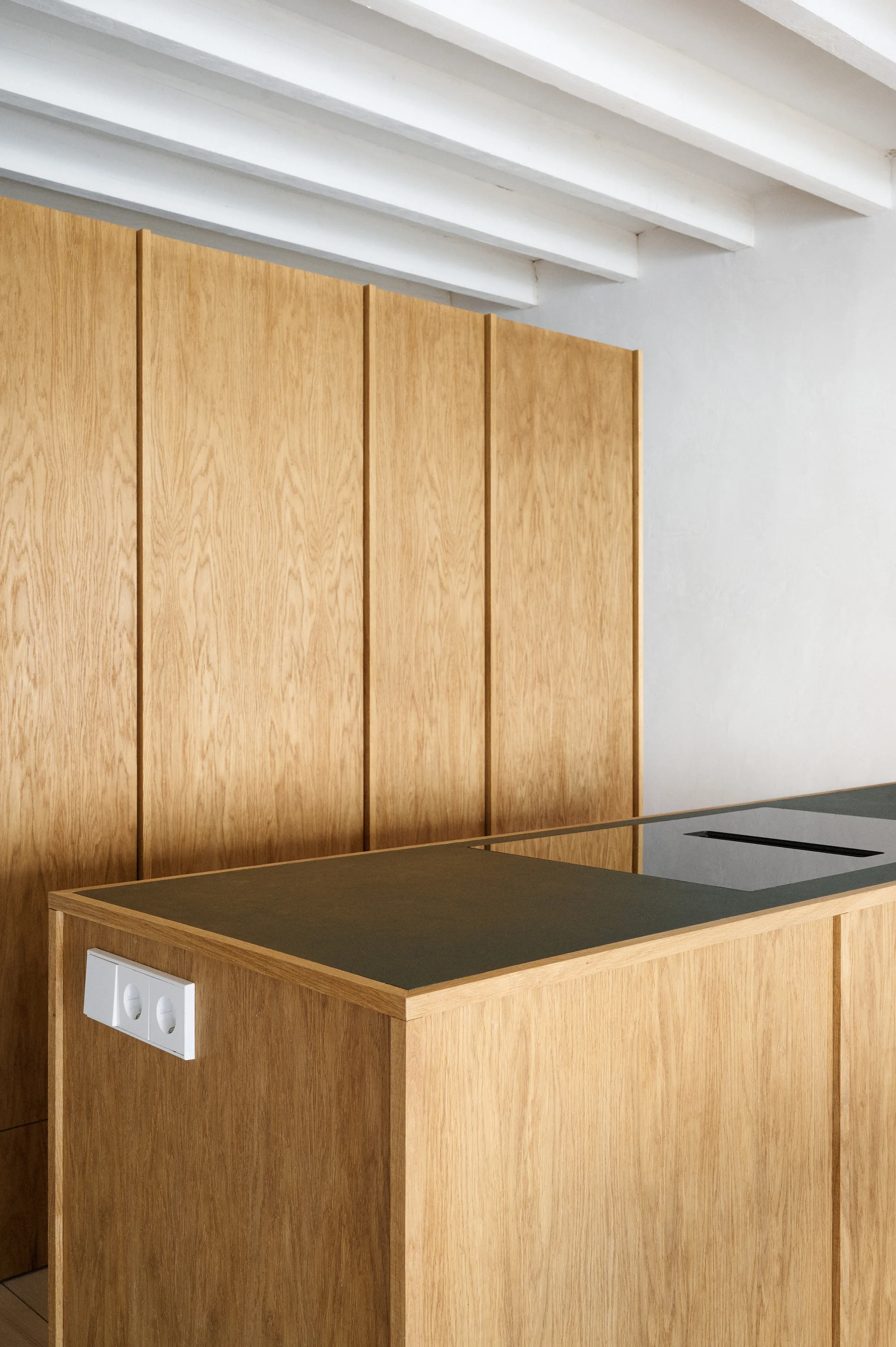 A minimalist kitchen features tall oak cabinets, a matching wood island with a dark gray countertop and integrated cooktop, and white ceiling beams.