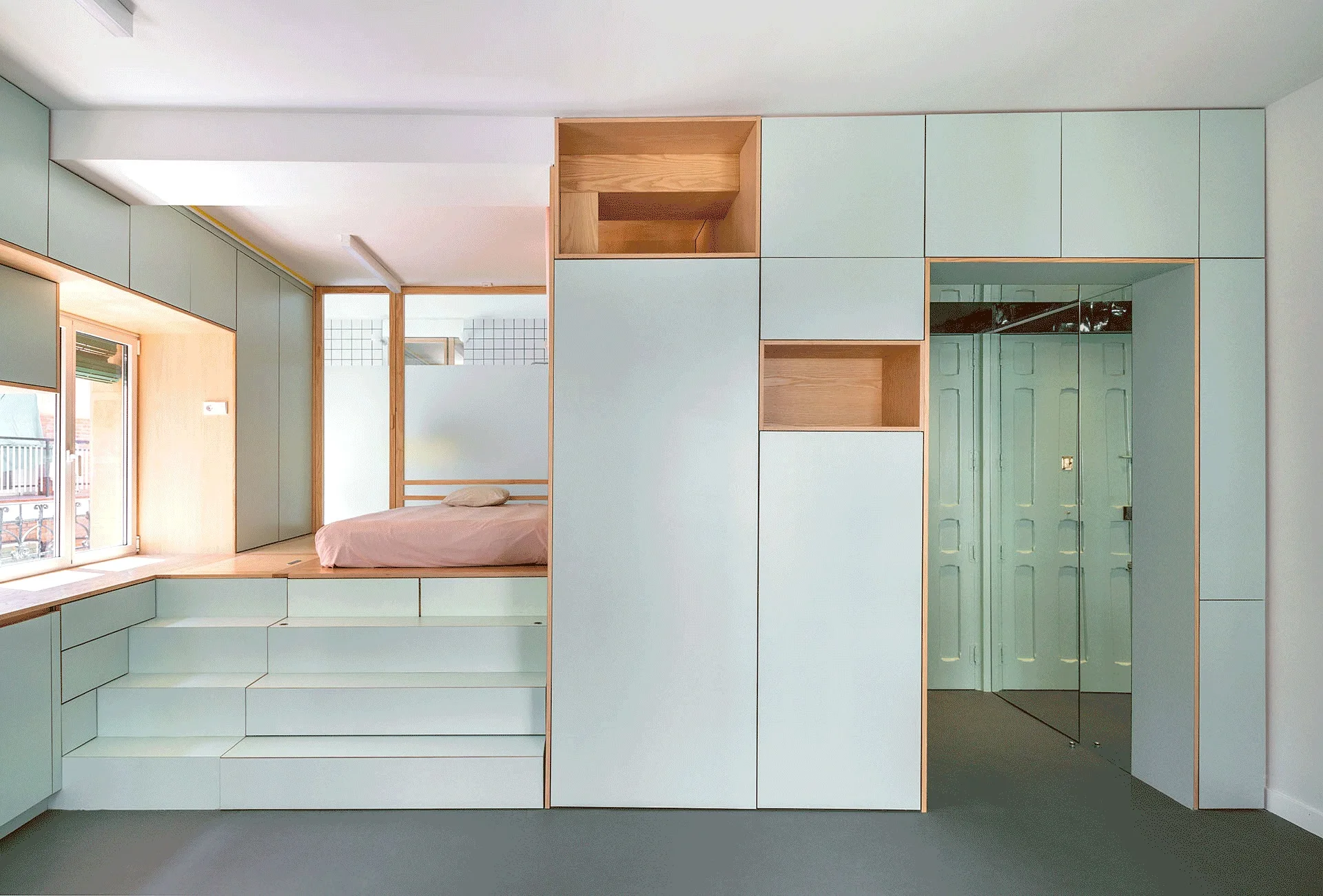 A custom raised bedroom platform with steps leading to a bed, bordered by floor-to-ceiling pale green built-in cabinetry and open wood niches.