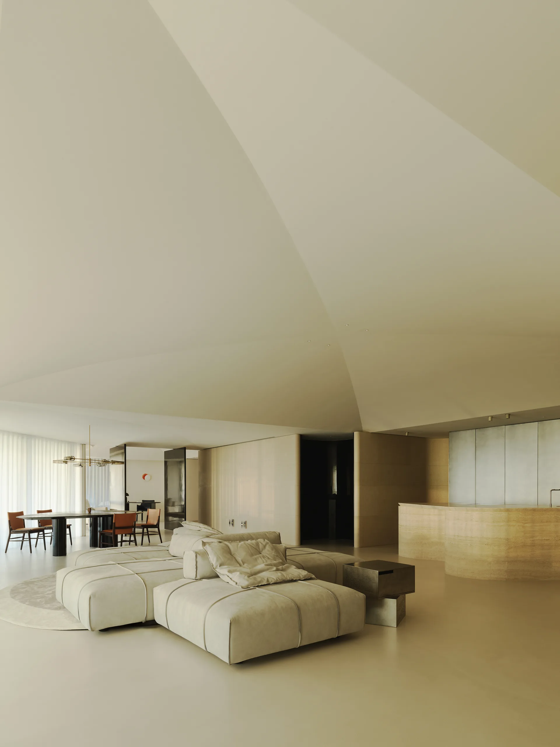 A spacious minimalist living room features a low-profile modular sectional sofa beneath an angular vaulted ceiling next to an open-plan kitchen and dining area.