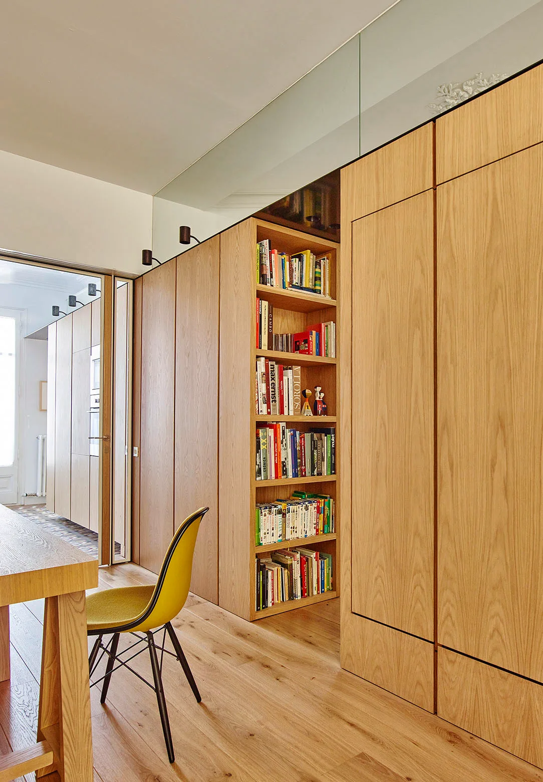 A modern interior features floor-to-ceiling light wood cabinetry with a built-in bookshelf, a yellow dining chair, and wood plank flooring.