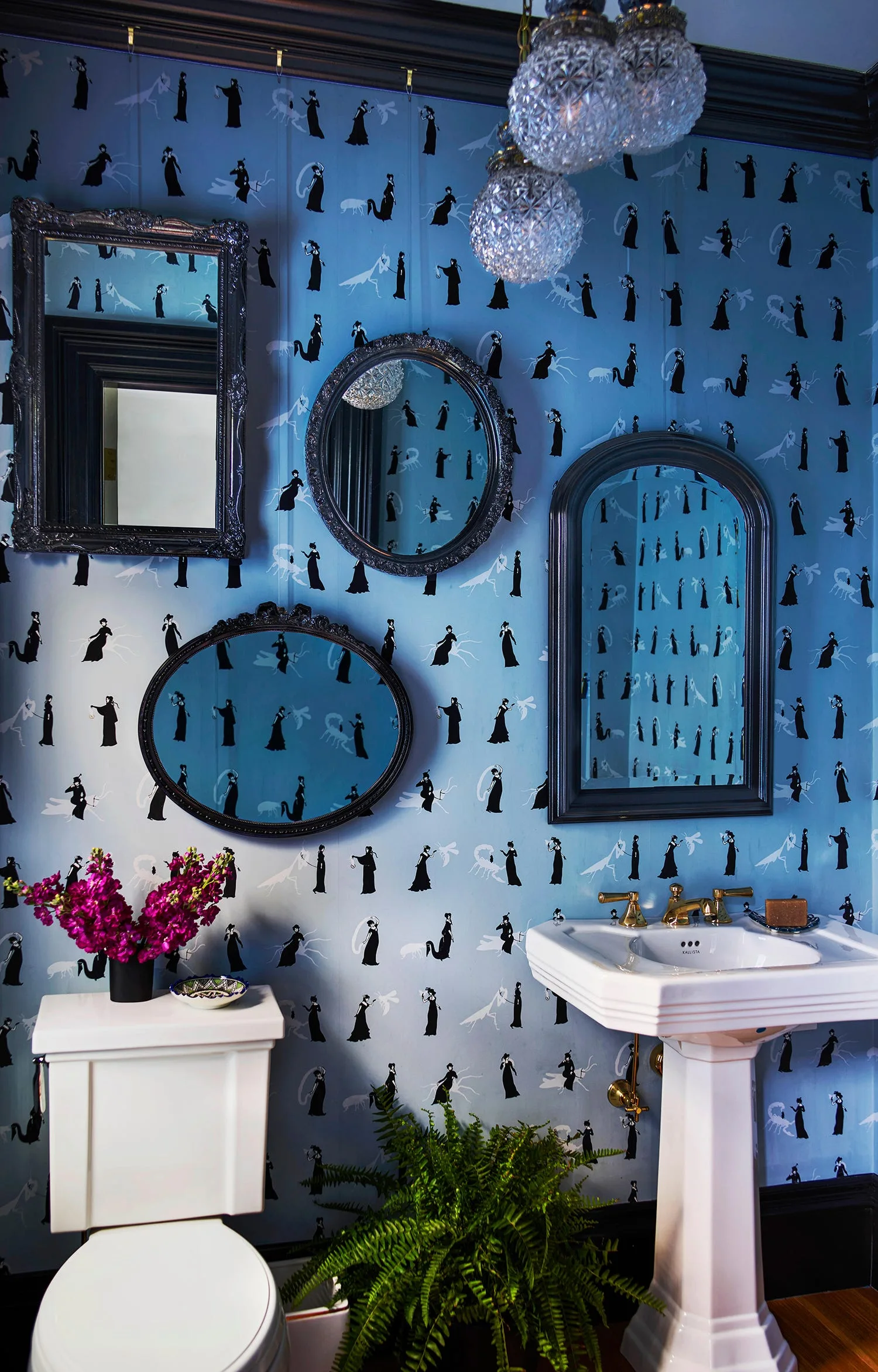 A powder room features blue graphic silhouette wallpaper, a white pedestal sink with brass fixtures, and a gallery of four dark-framed mirrors.
