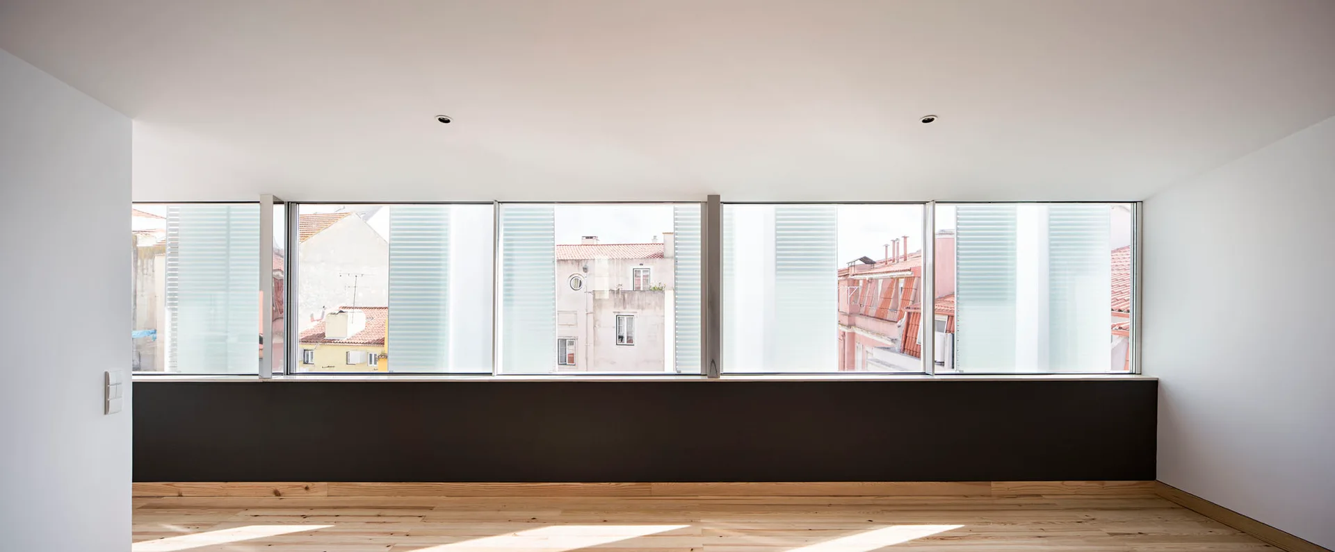 An empty minimalist room featuring a long horizontal window wall above a dark gray panel, white walls, and light wood plank flooring.