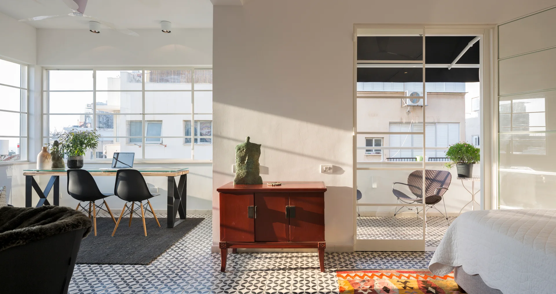 A bright room with geometric floor tiles features a wooden cabinet, a glass-top desk with black chairs, and a glass door opening to a balcony.