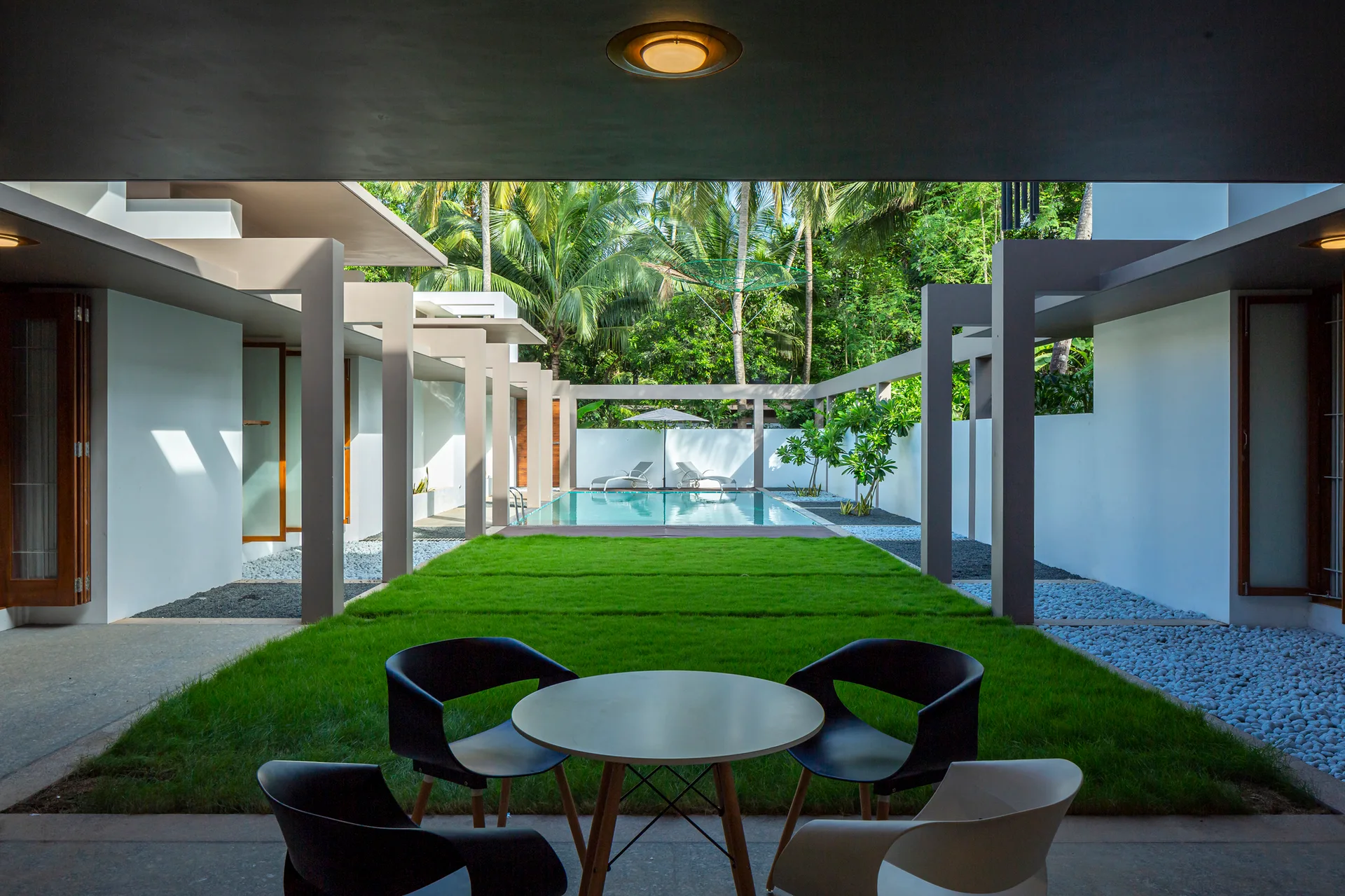 An outdoor courtyard with a central green grass lawn, geometric concrete pillars, patio furniture, and a swimming pool.