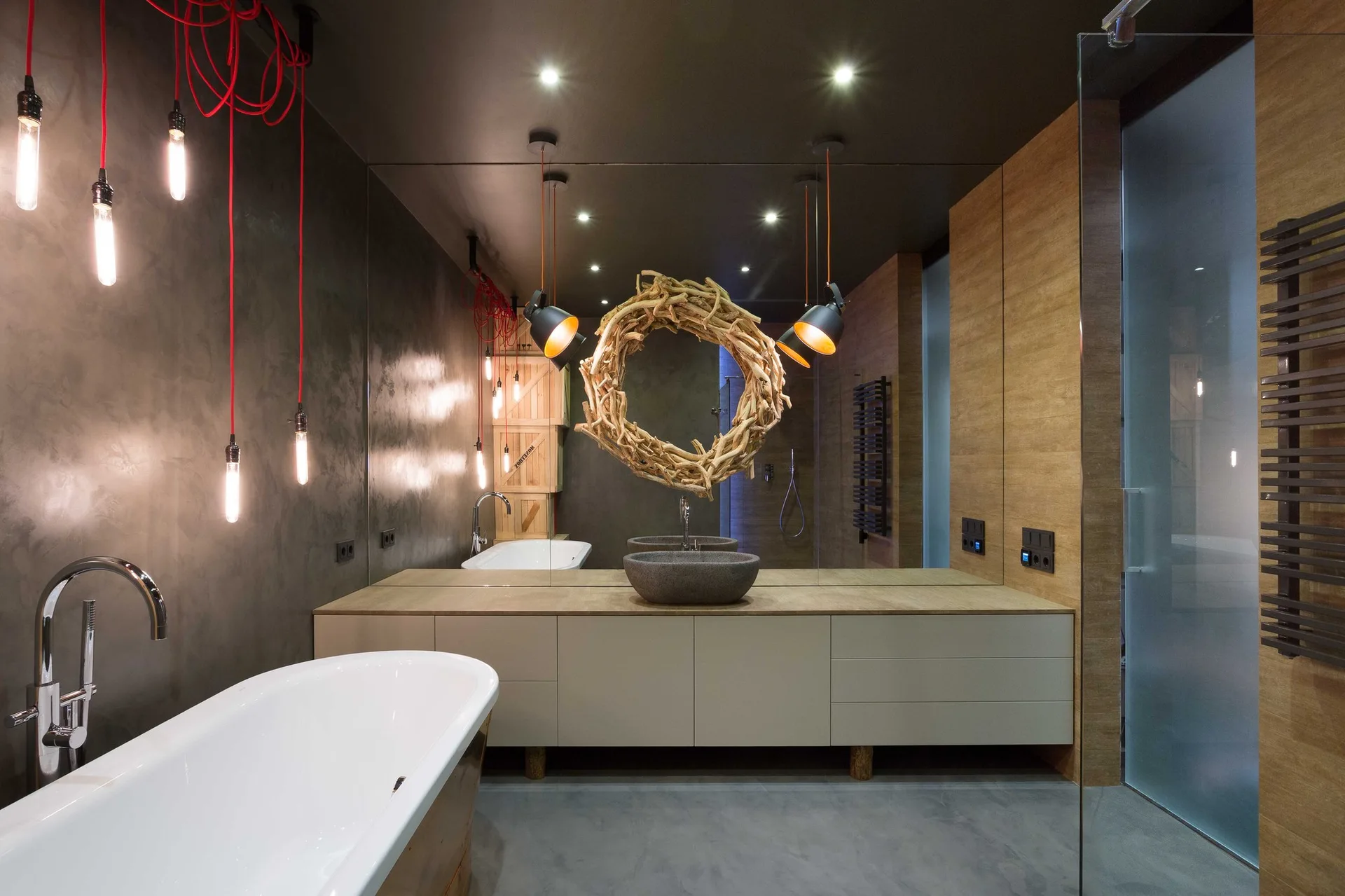 A dark gray bathroom featuring a freestanding white bathtub, a floating vanity with a stone vessel sink, and a circular driftwood sculpture on the mirror.