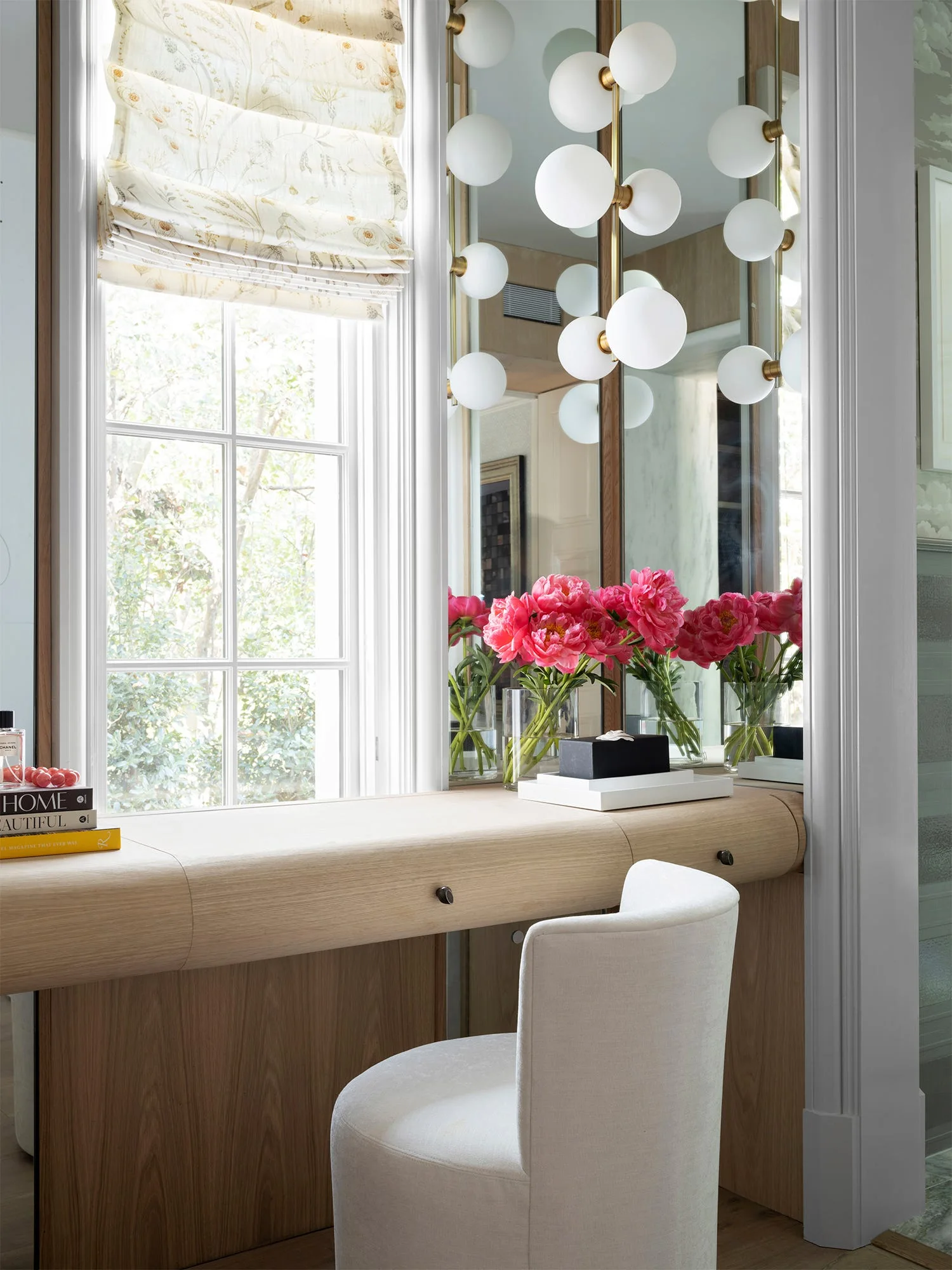 A light wood built-in dressing vanity in an alcove next to a window features a white upholstered chair, pink peonies, and a multi-globe pendant light.