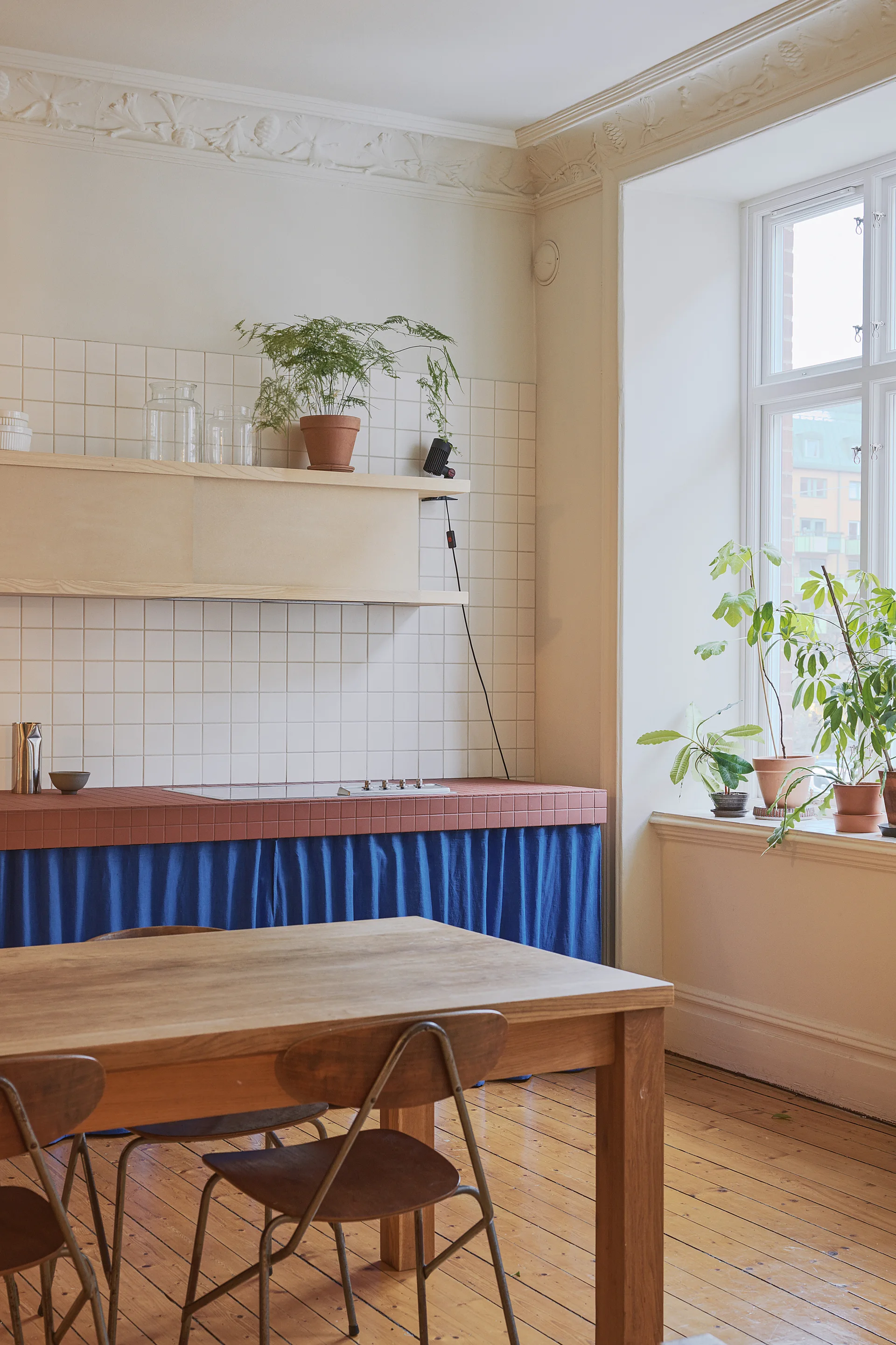 A kitchen featuring a terracotta-tiled countertop with a blue fabric skirt, a wooden dining table, a white tiled backsplash, and a large window.