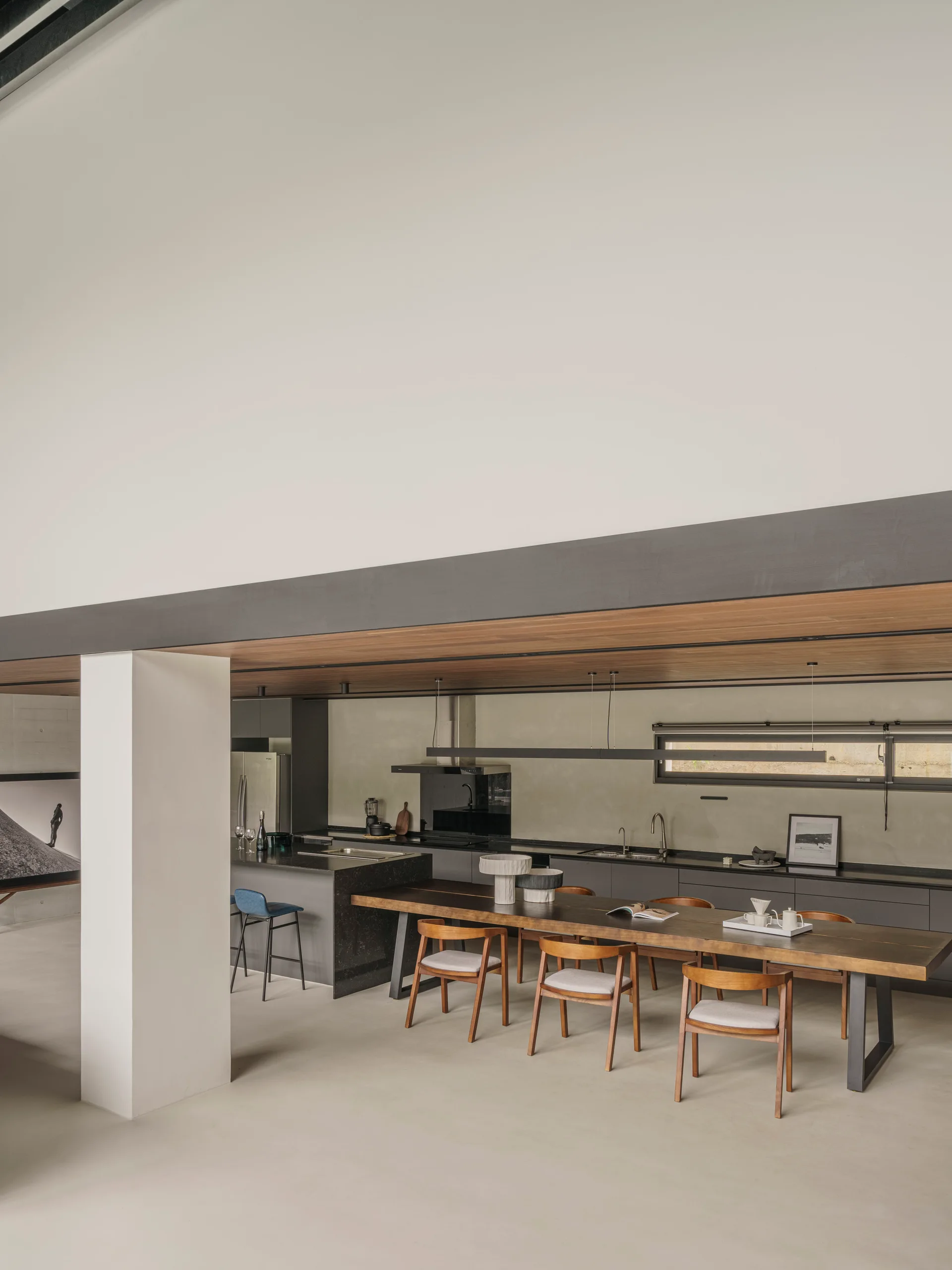 A minimalist open-plan kitchen and dining area with a long wooden table, a dark stone island, and a prominent dark horizontal ceiling beam.