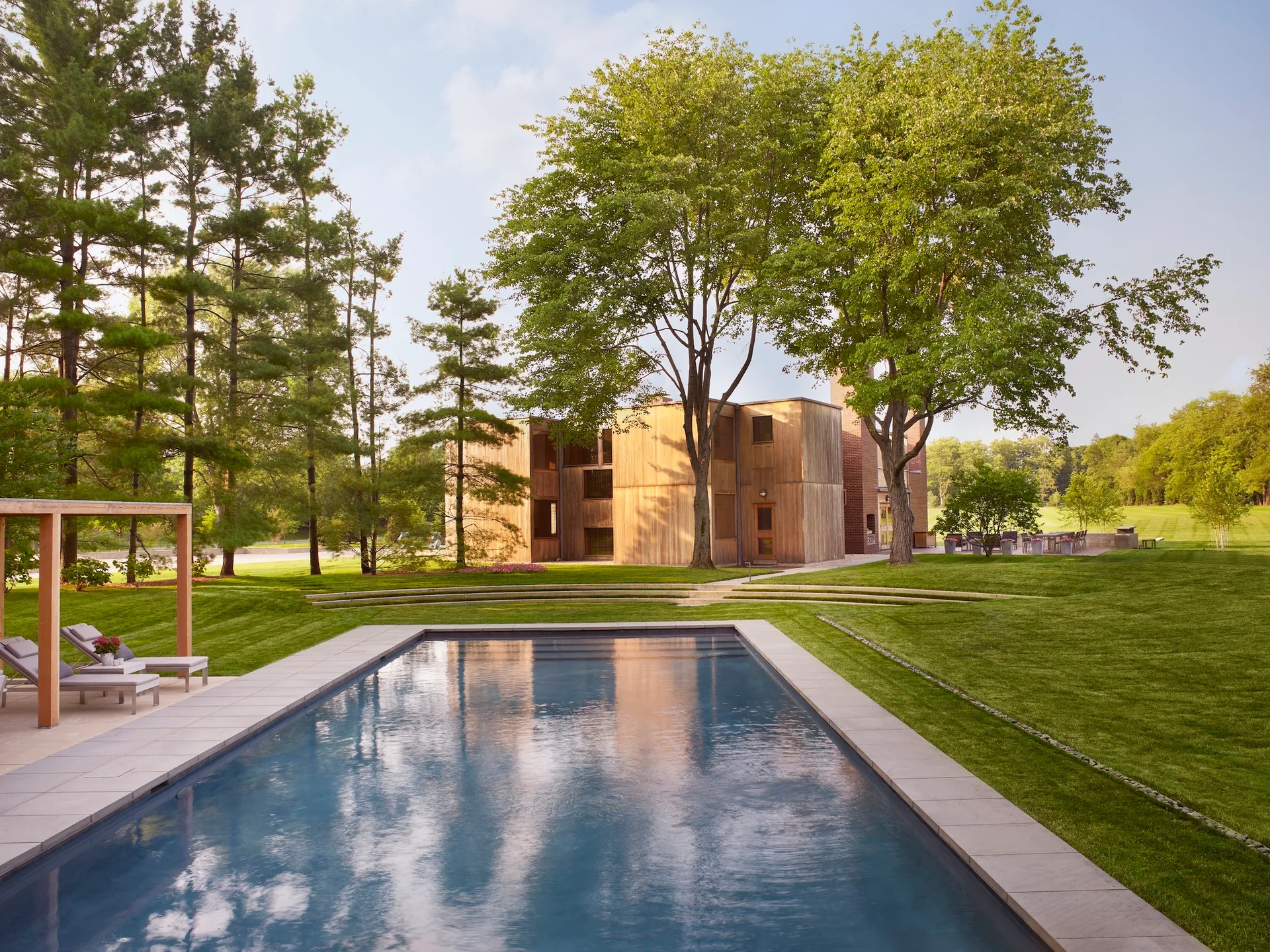 A rectangular swimming pool is set within a vast lawn leading to a modernist wood-clad house bordered by tall green trees.