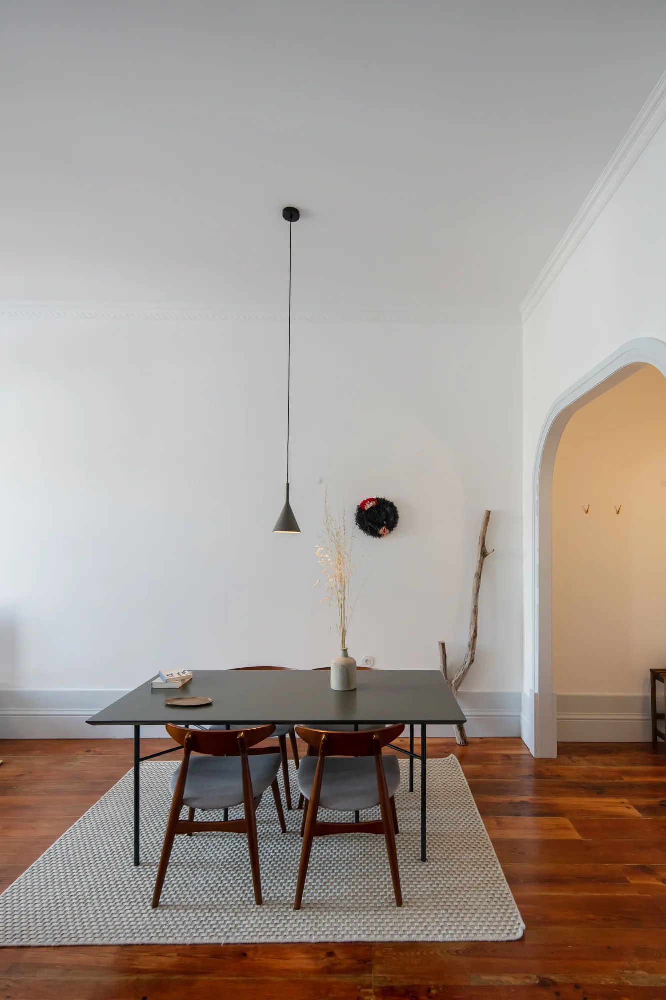 A black dining table with wooden chairs sits on a woven rug beneath a black pendant light in a white room with an arched doorway.