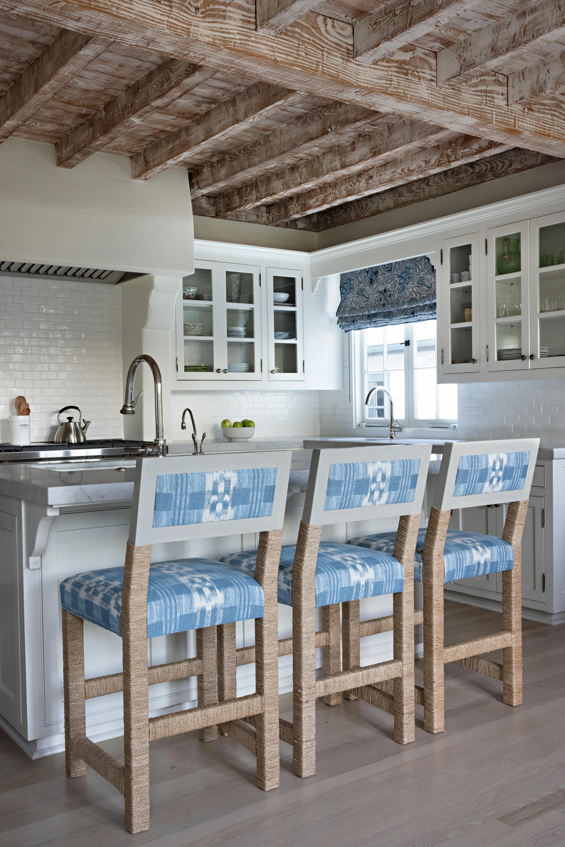 Three rope-wrapped bar stools with blue fabric sit at a white kitchen island under a rustic wood-beamed ceiling.