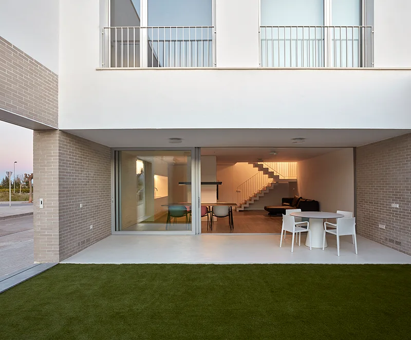 A modern patio features a white circular dining set next to a lawn, with large sliding glass doors revealing an open-concept interior and staircase.
