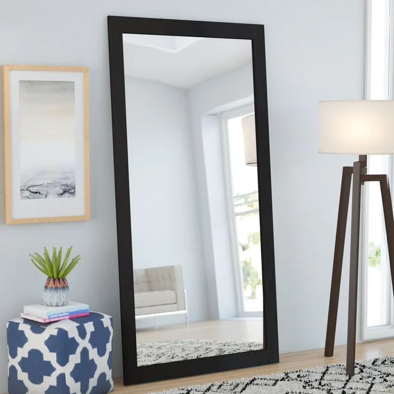 A dark-framed full-length mirror leans against a light gray wall next to a tripod floor lamp and a blue patterned cube ottoman.