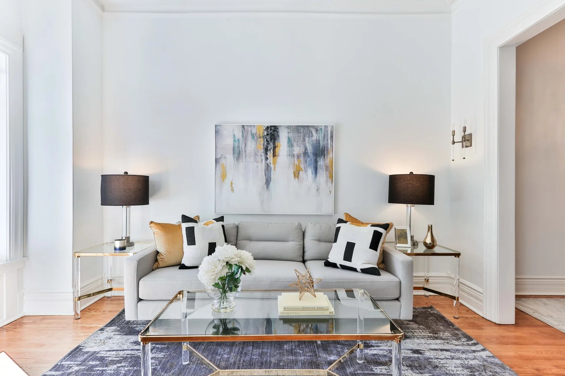 A bright white living room with a symmetrical arrangement of a light gray sofa flanked by glass end tables, black-shaded lamps, and abstract wall art.