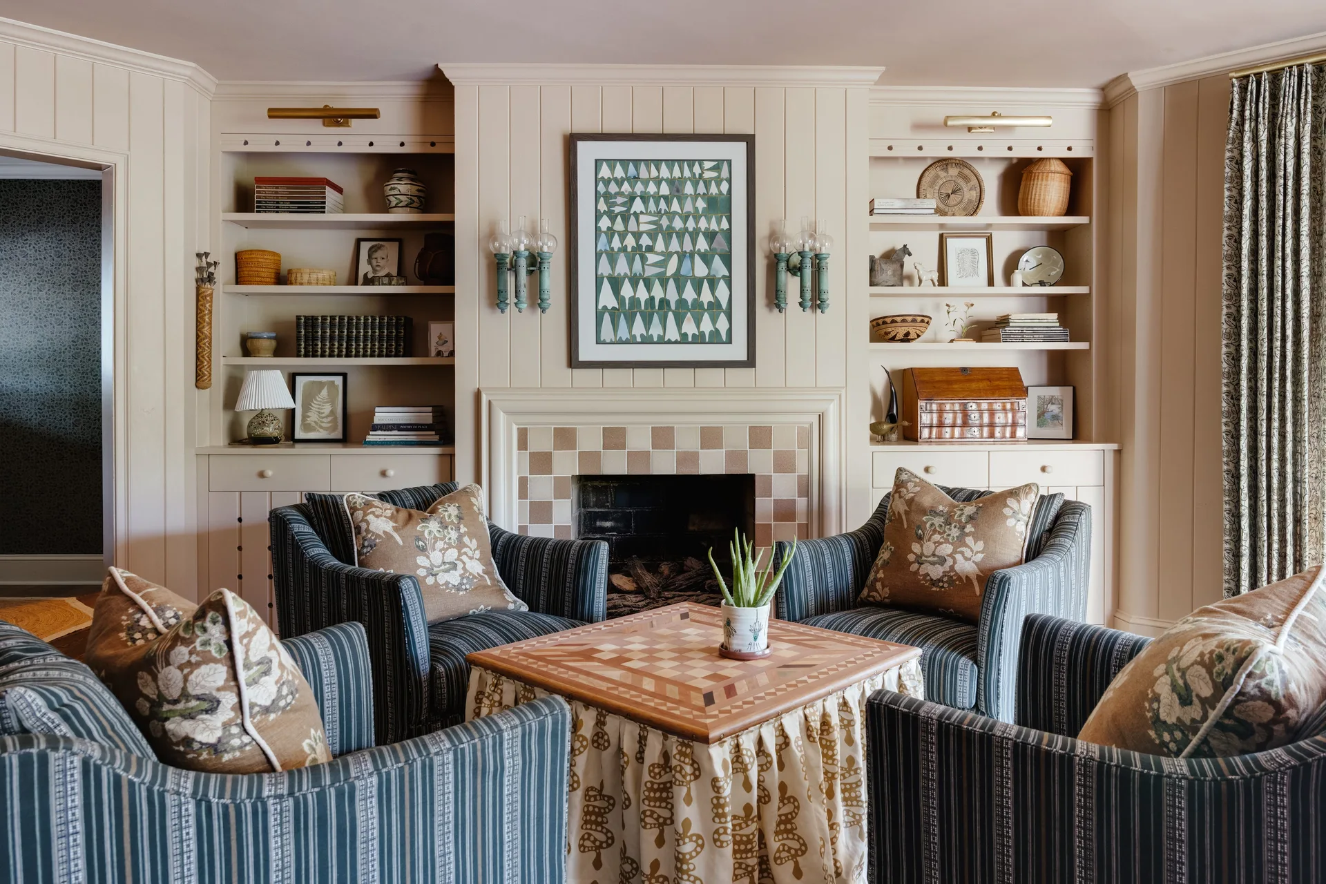 Four blue striped armchairs surround a skirted wooden game table in front of a tiled fireplace flanked by symmetrical built-in bookshelves.