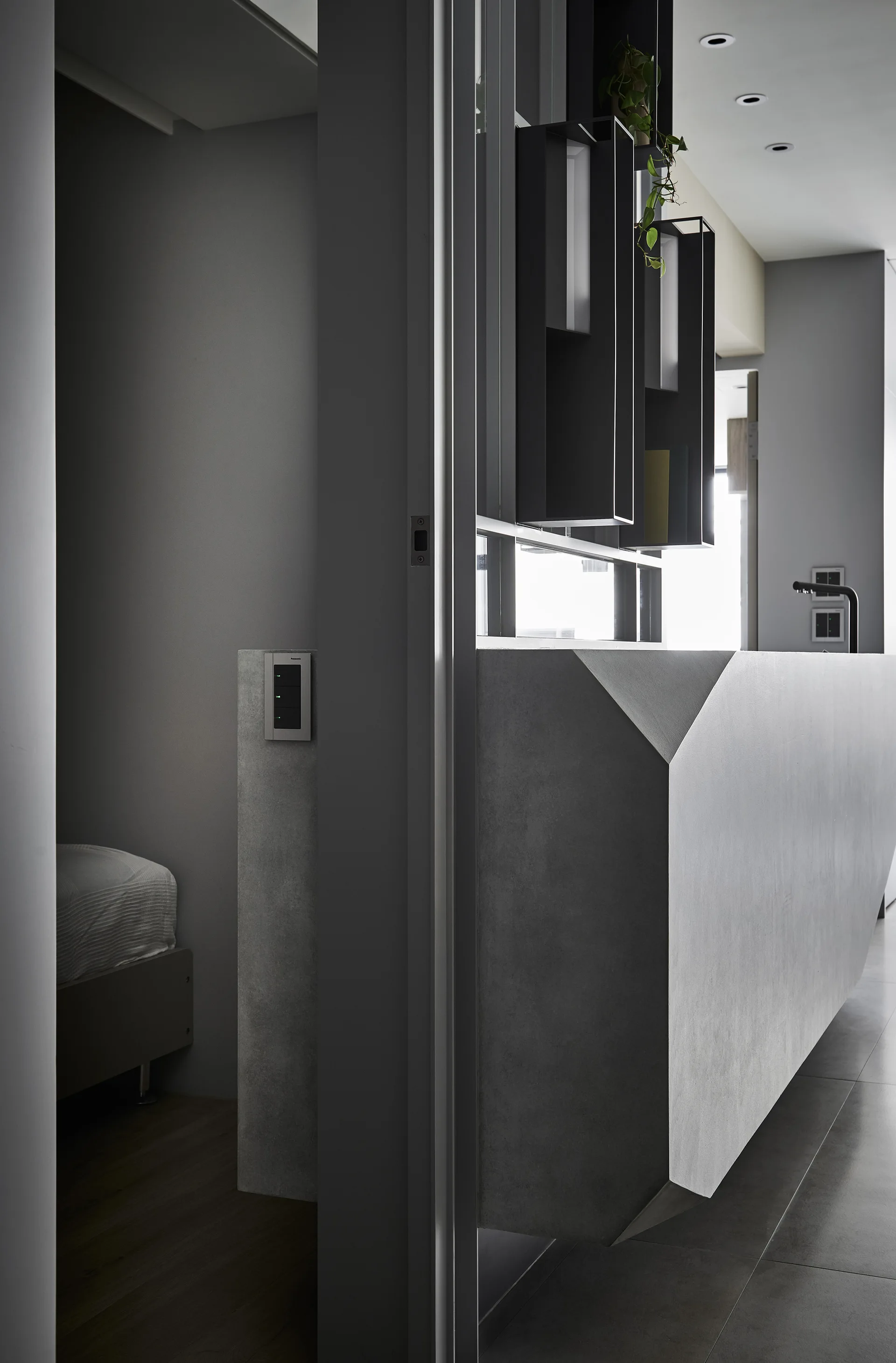 A faceted grey concrete partition wall stands in a minimalist apartment, featuring geometric angles, black open shelving, and a view into a bedroom.