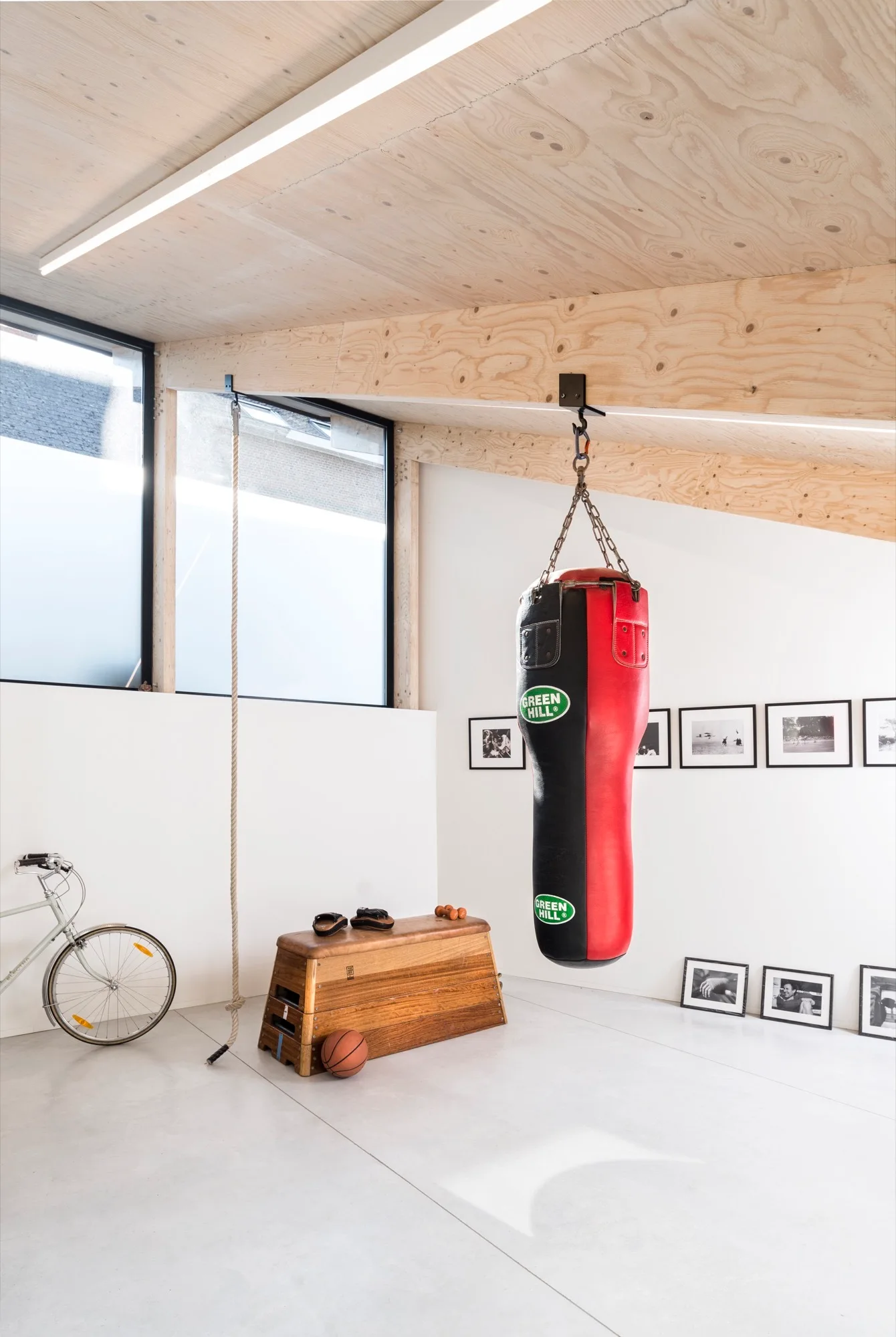 A minimalist home gym featuring a red and black punching bag hanging from a plywood ceiling beam, next to a wooden gym bench and a climbing rope.