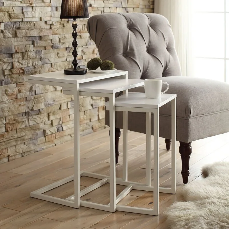 Three white metal nesting tables of varying heights stand beside a tufted grey armchair on wood floors in front of a stacked stone wall.