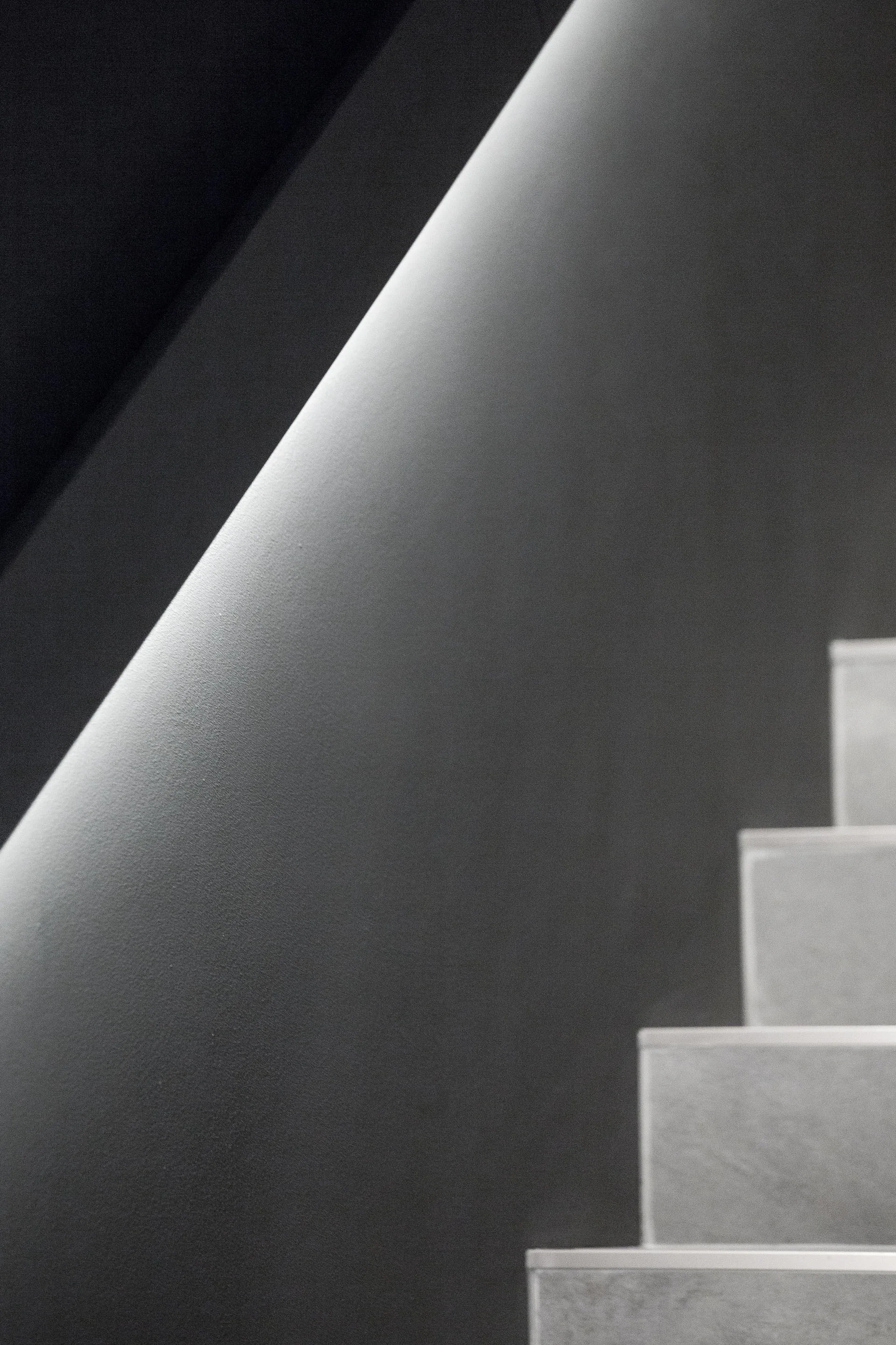 A minimalist grey staircase with stone-like treads rises next to a dark grey wall illuminated by a bright, diagonal strip of recessed linear lighting.