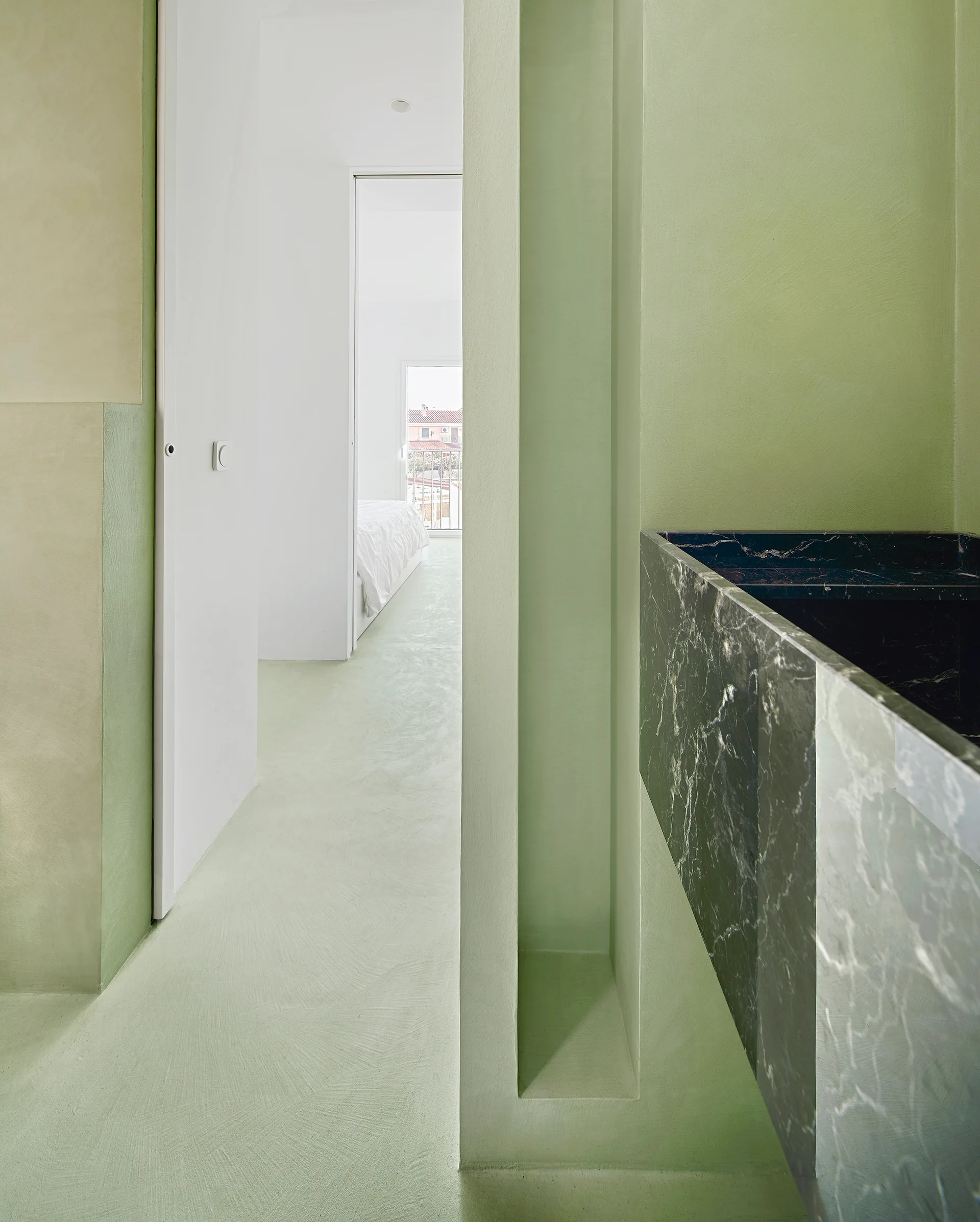 A sage green hallway with plastered walls and floors features a vertical wall niche and a black veined marble sink basin leading to a white bedroom.