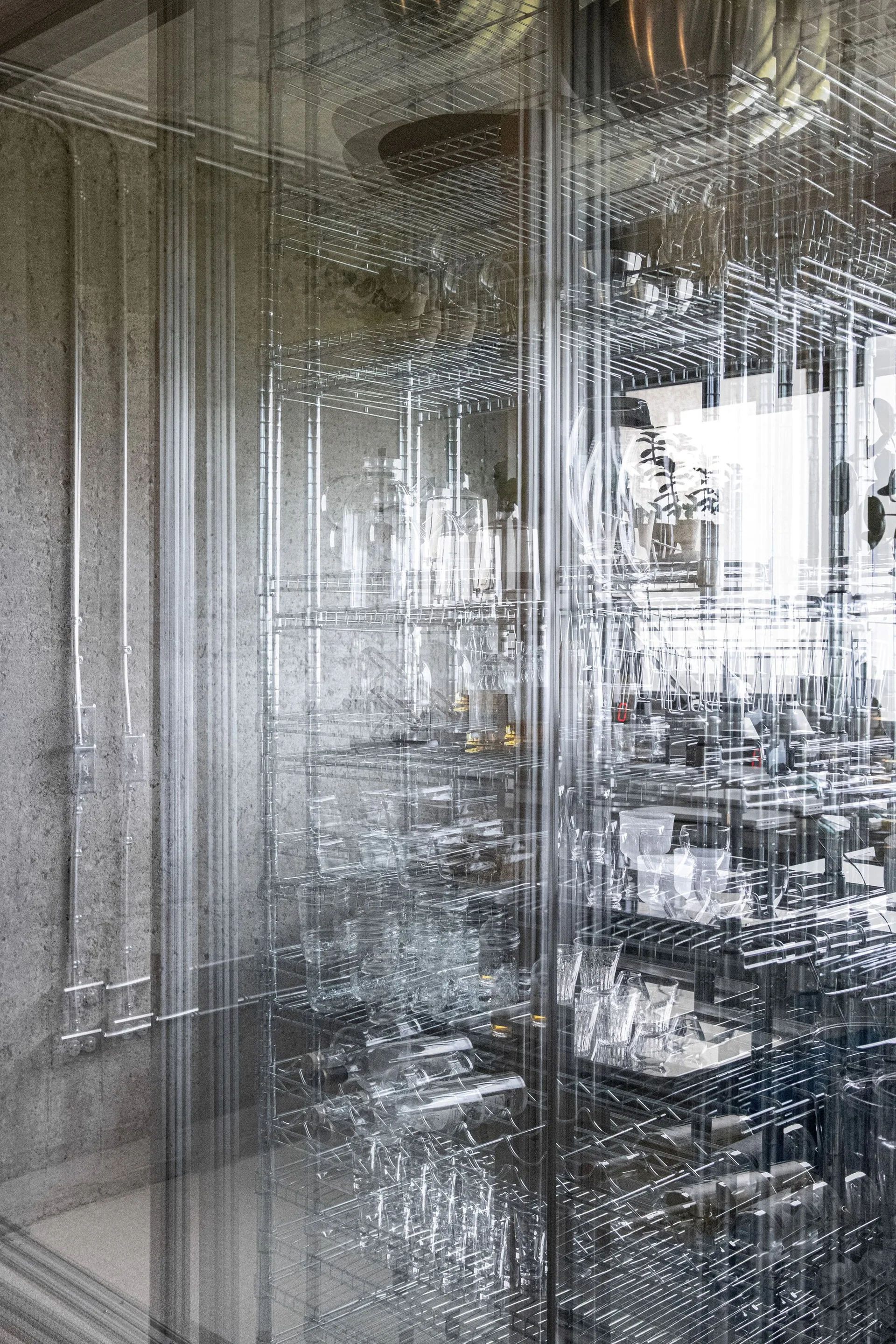 A metal wire shelving unit displaying glassware is seen through a reflective glass partition against a concrete wall.