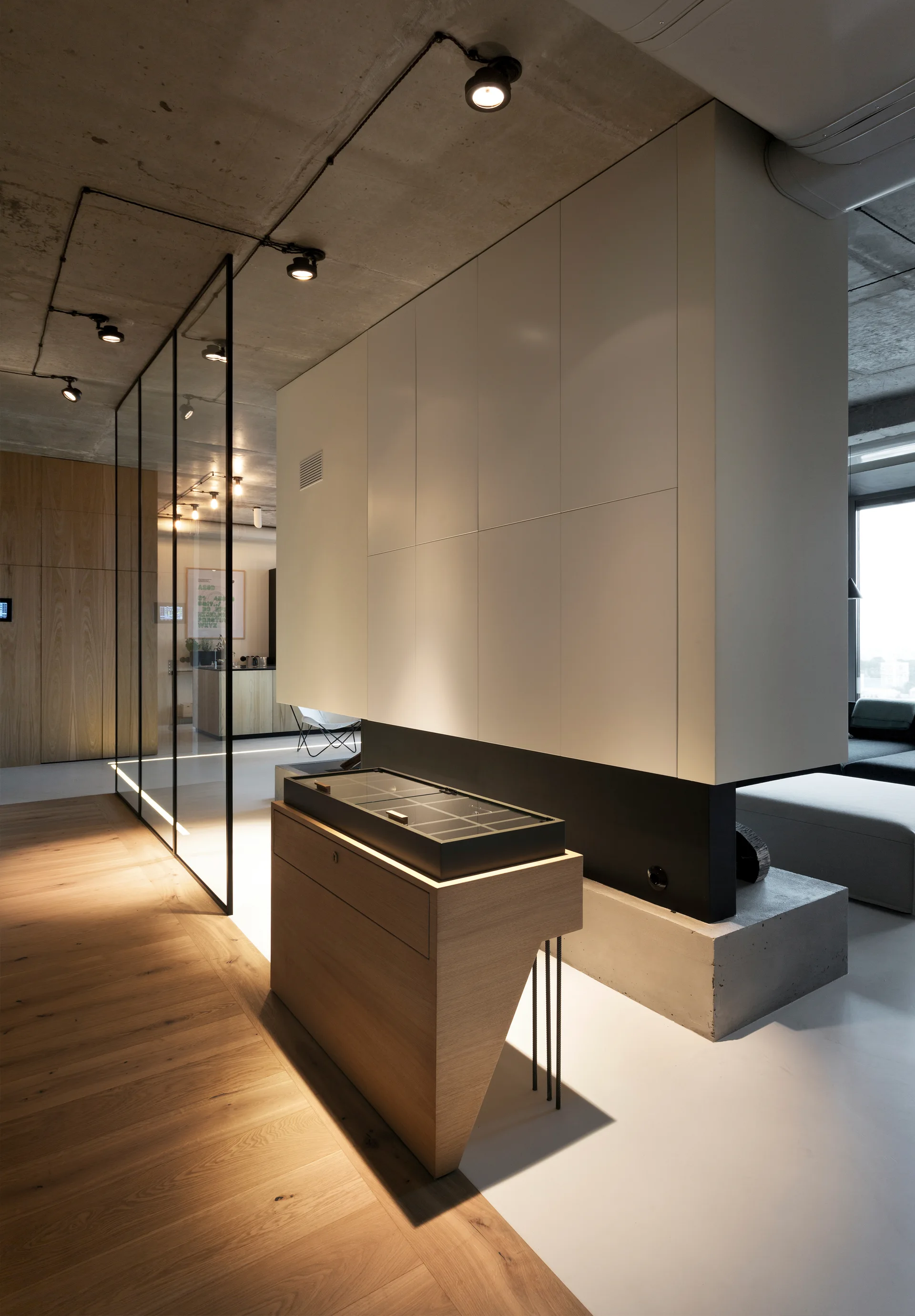 A modern minimalist interior with an exposed concrete ceiling, a large white storage wall, a glass partition, and a light wood console table.