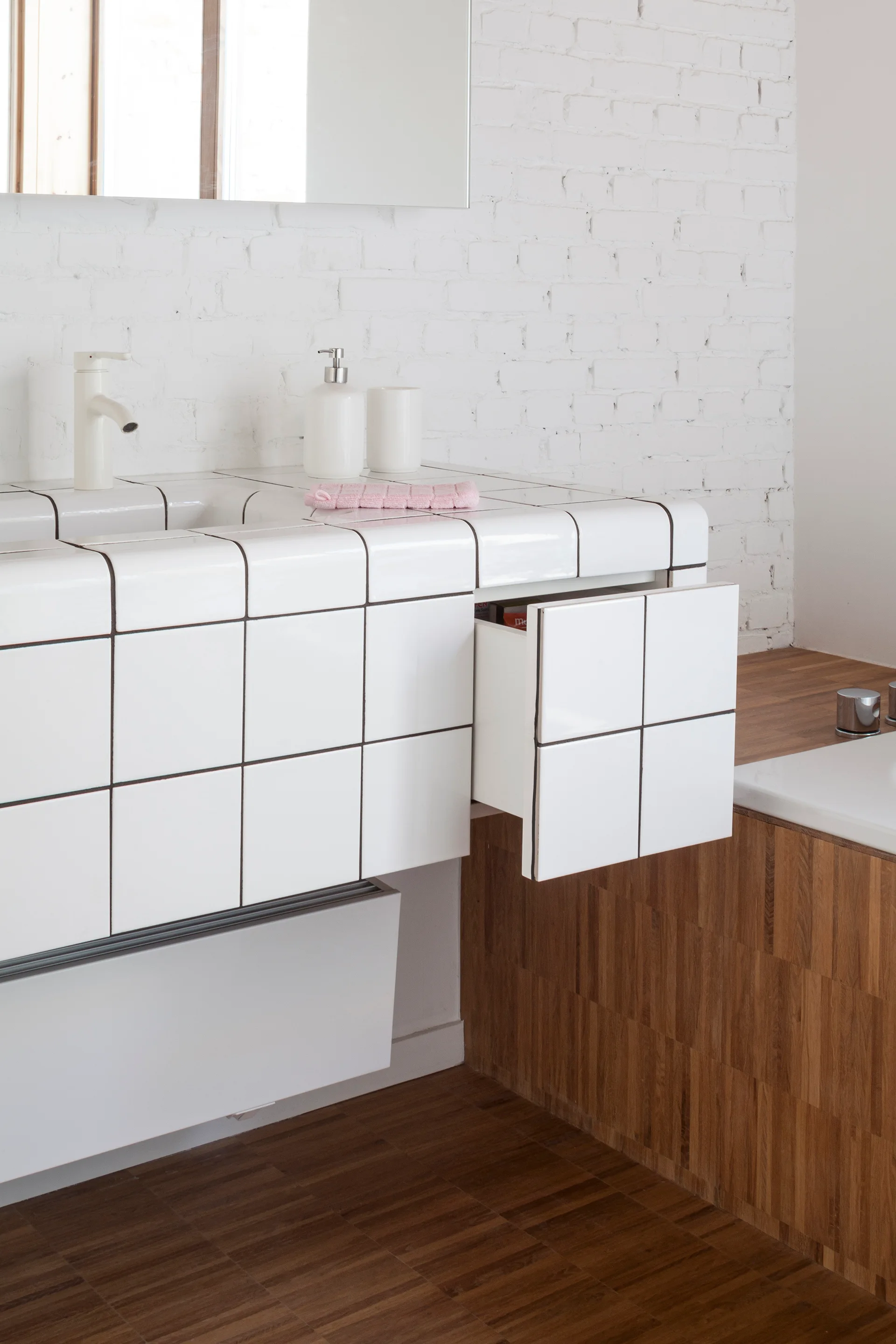 A modern bathroom vanity wrapped in white square tiles with dark grout features an open drawer against a white brick wall.